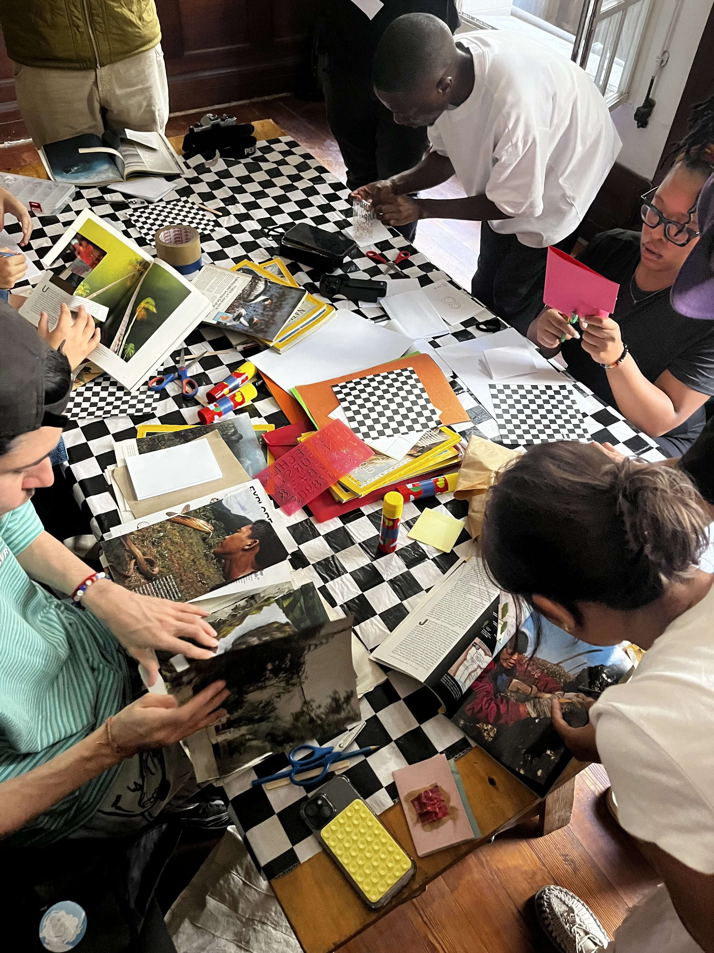 People gathered around a table covered with a black-and-white checkered tablecloth, engaging in arts and crafts activities with magazines, scissors, glue, and paper. Some individuals are looking through magazines, while others are cutting or assembli
