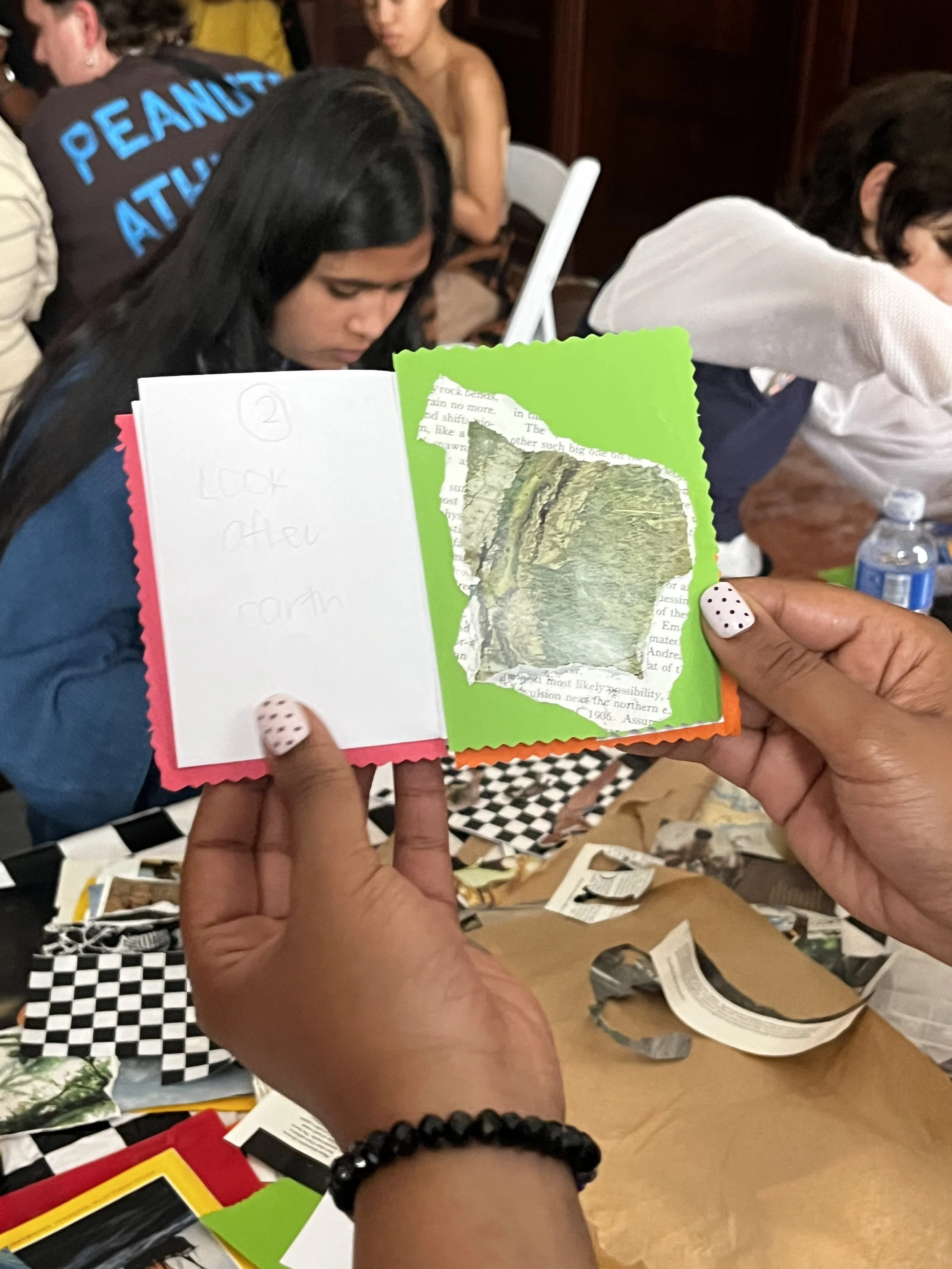 A person holding a colorful handmade card featuring a torn paper landscape. The card has a white, pink, green, and orange border, with text on the inside. In the background, several people are seated at a table, possibly at an indoor gathering or eve