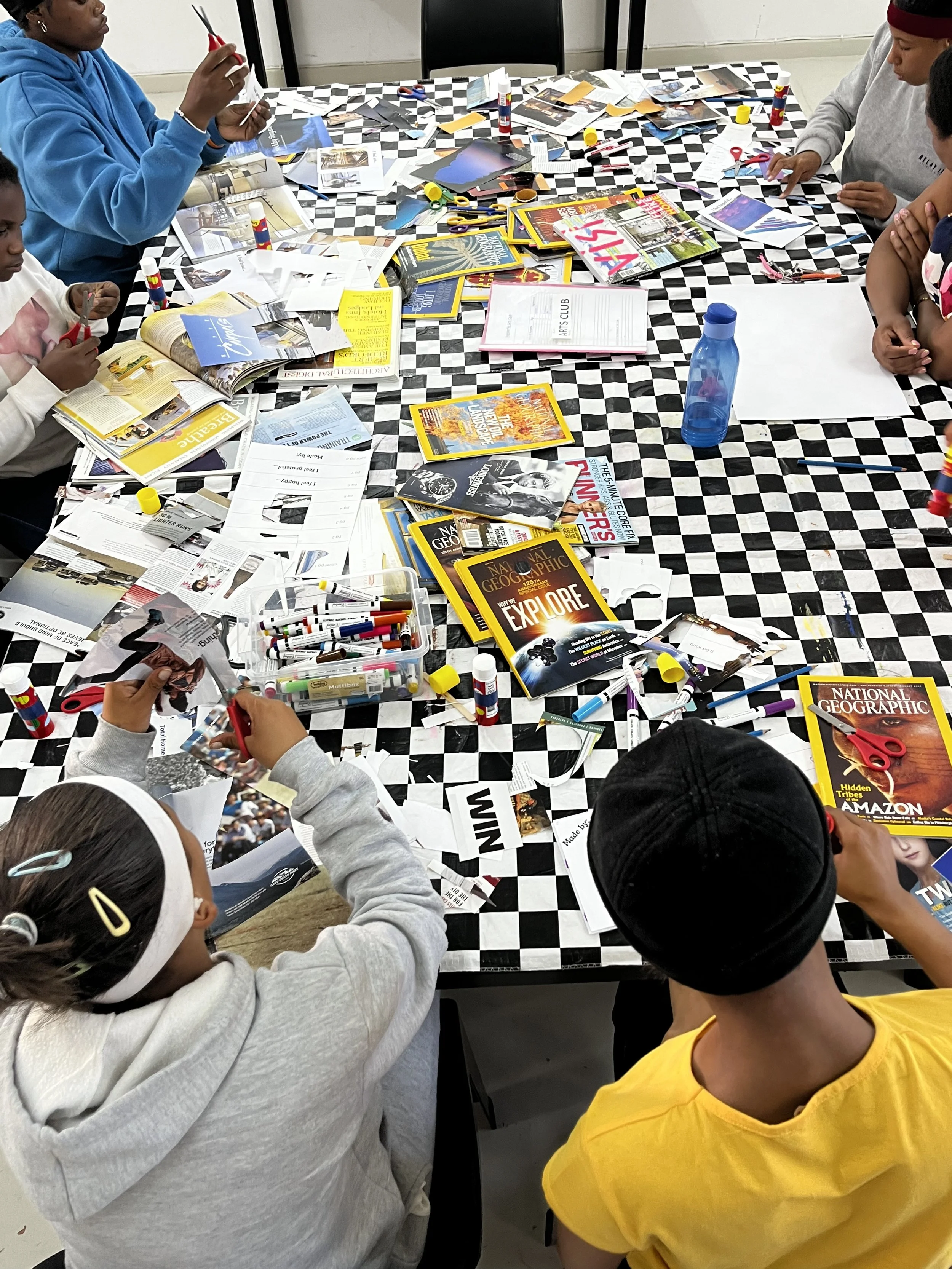 Children working on arts and crafts projects at a table covered with magazines, scissors, glue, and paper in a classroom or activity space.