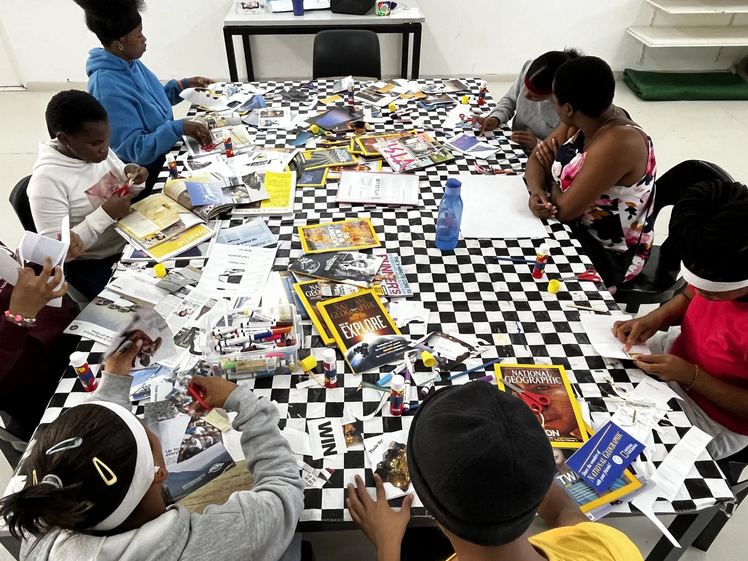 Children and adults gathered around a table covered with magazines, pamphlets, scissors, glue sticks, and markers, working on a collage or craft project.