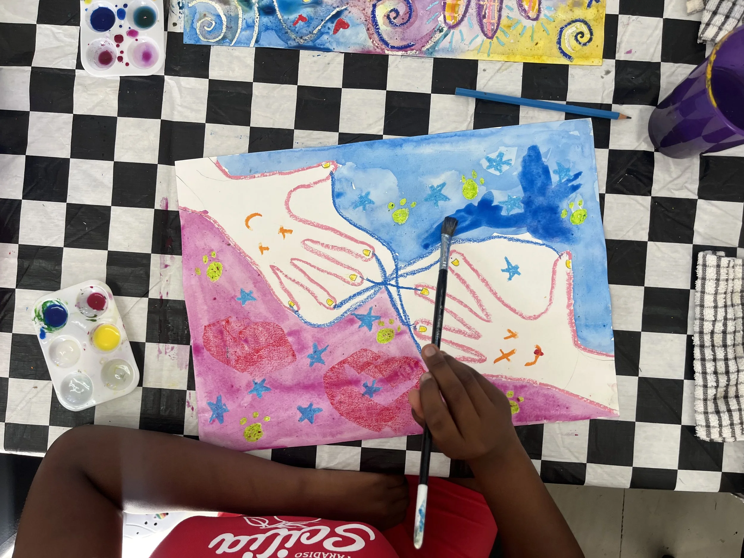 Child painting colorful artwork with star and swirl designs using watercolors on large paper, and palette of paints nearby on checkered tablecloth.