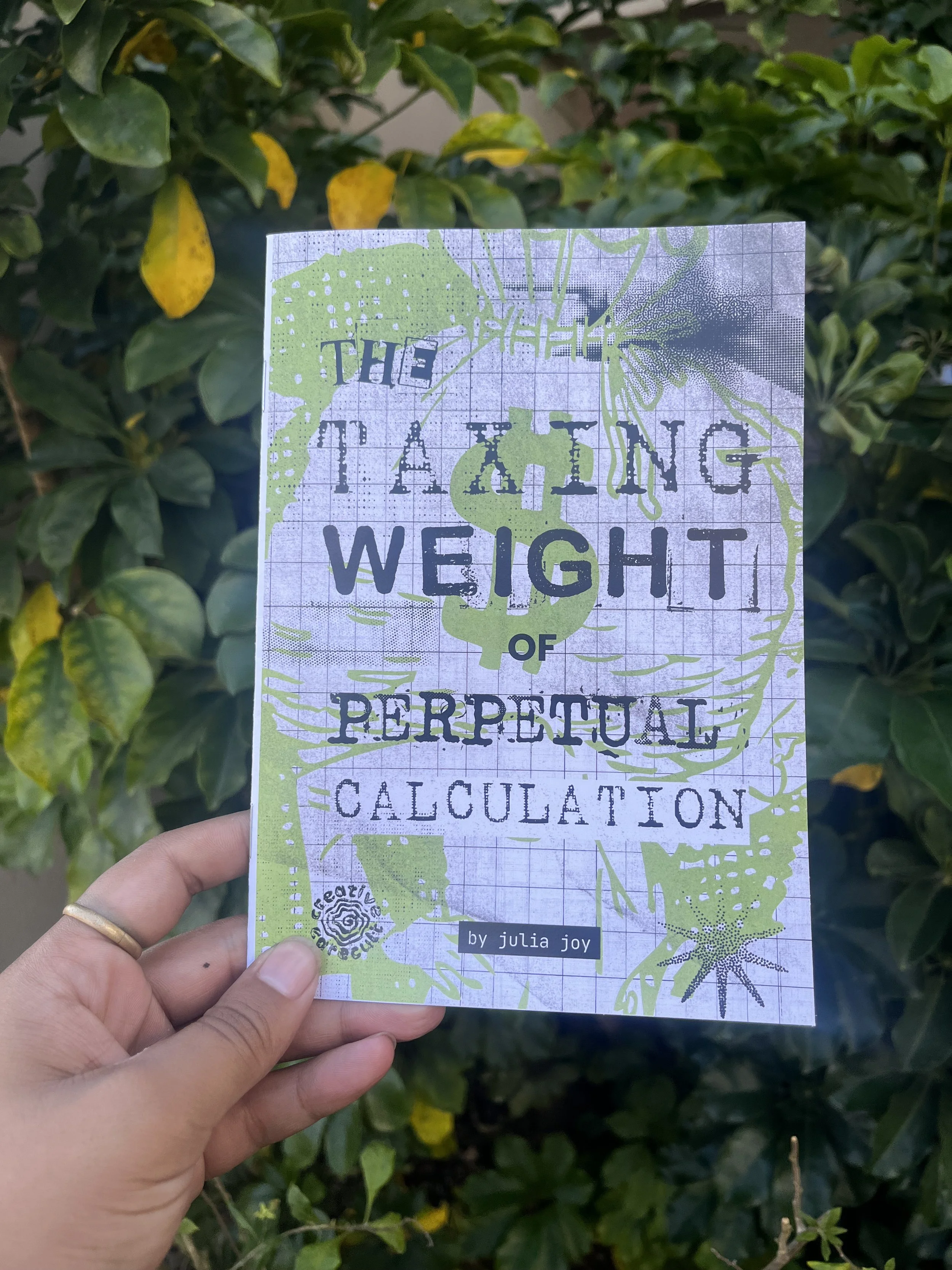 A hand holding a book titled 'The Teaching of Perpetual Calculation' by Julia Joy, held in front of green leafy bushes with some yellowing leaves.