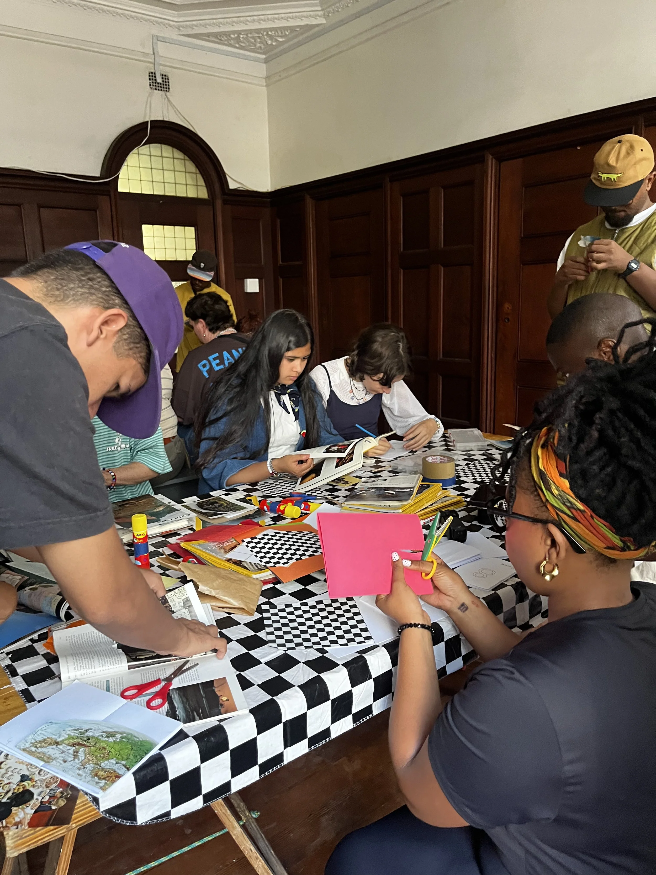 People gathered around a checkered table filled with books, magazines, scissors, and craft supplies, working on a creative project in a room with wood-paneled walls.