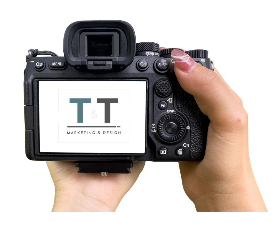 A person holding a DSLR camera displaying a logo for T & T Marketing & Design on its screen.