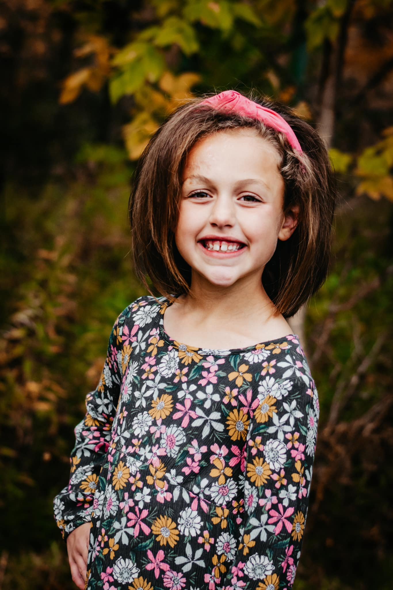 A young girl with a pink headband, shoulder-length brown hair, and a big smile shows her missing a front tooth. She is wearing a long-sleeved floral dress with pink, white, and yellow flowers. The background features foliage in shades of green and ye