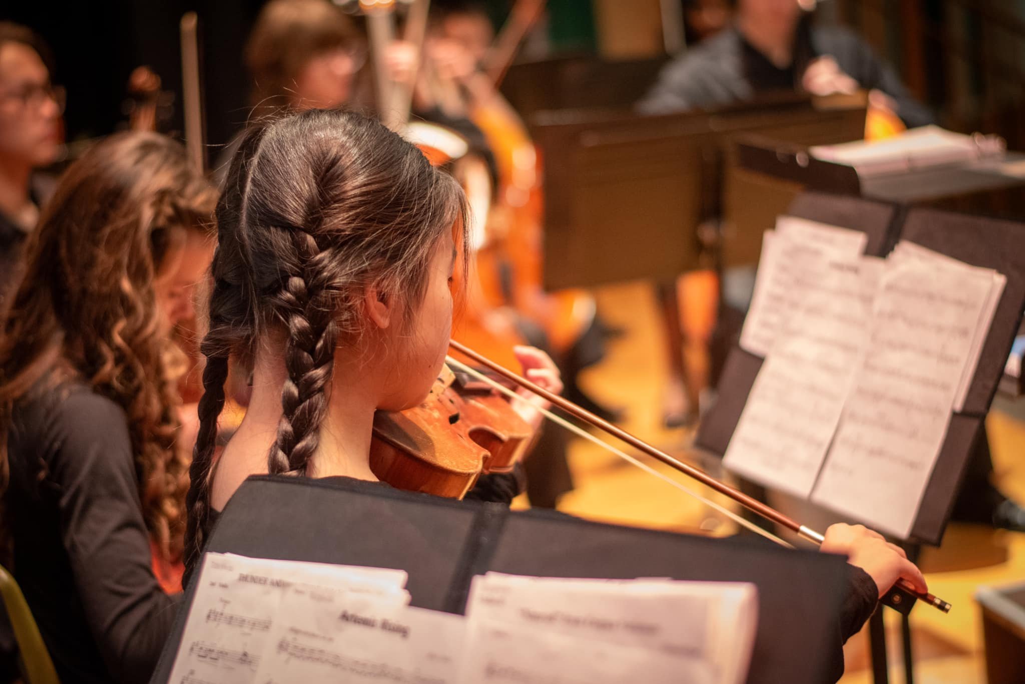Young woman with braided hair playing the violin during an orchestra performance, with sheet music on stand in front of her and other musicians in the background.