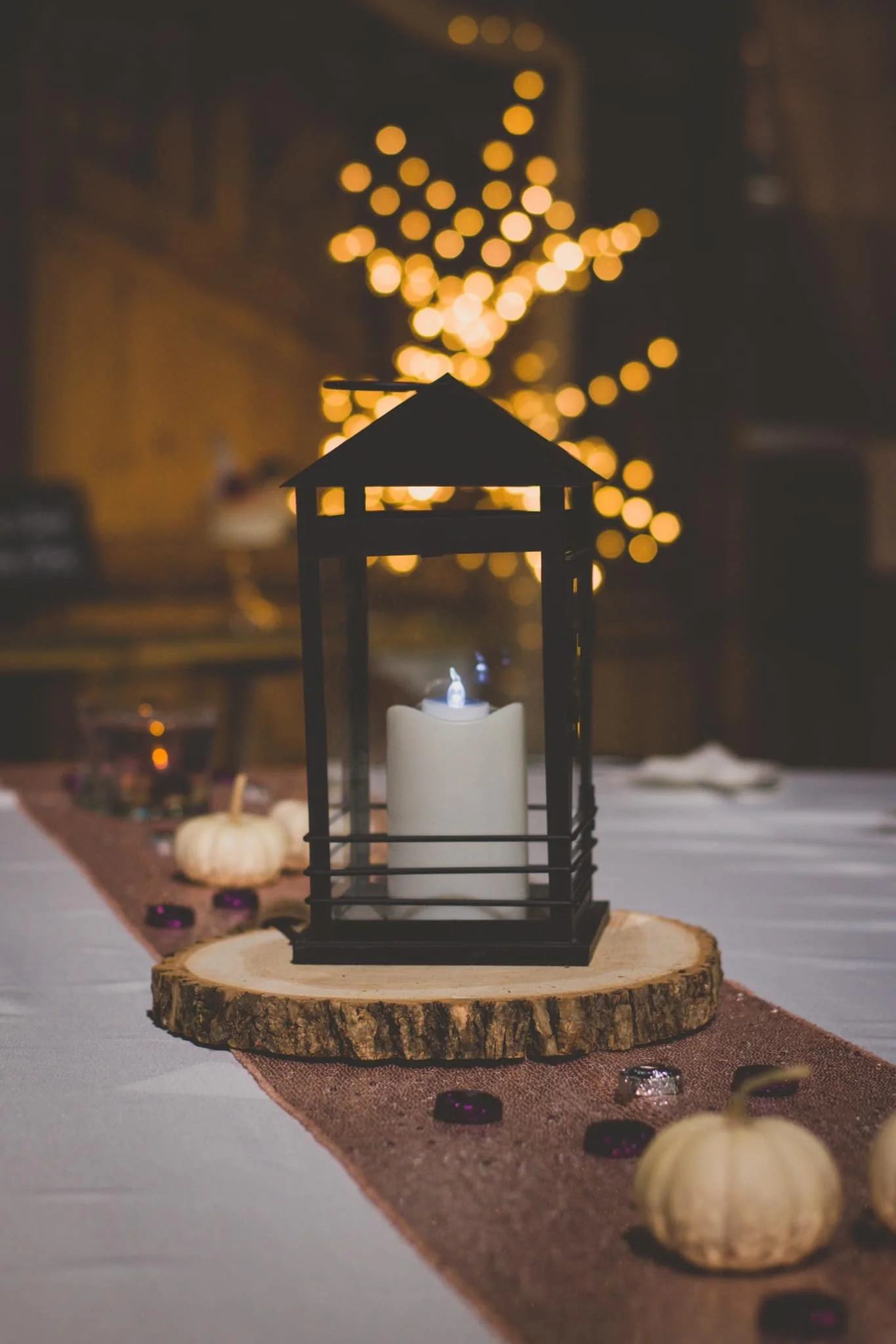 A table centerpiece with a black lantern containing a white candle on a round wooden slice. In the background, blurred yellow fairy lights create a warm glow. Small pumpkins and purple and clear gems decorate the table runner.