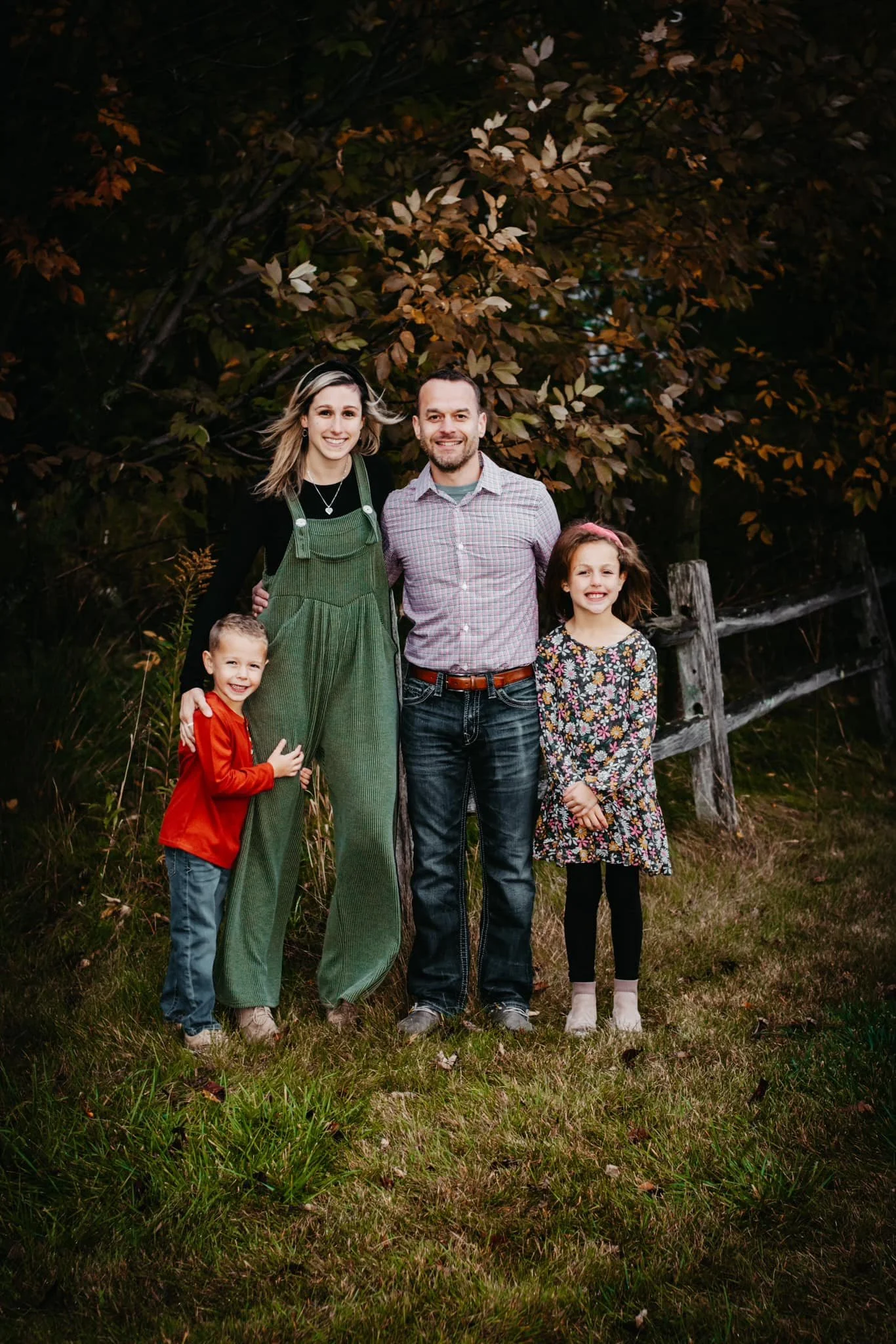 Family of five standing outdoors on grass with autumn leaves in the background, smiling at the camera