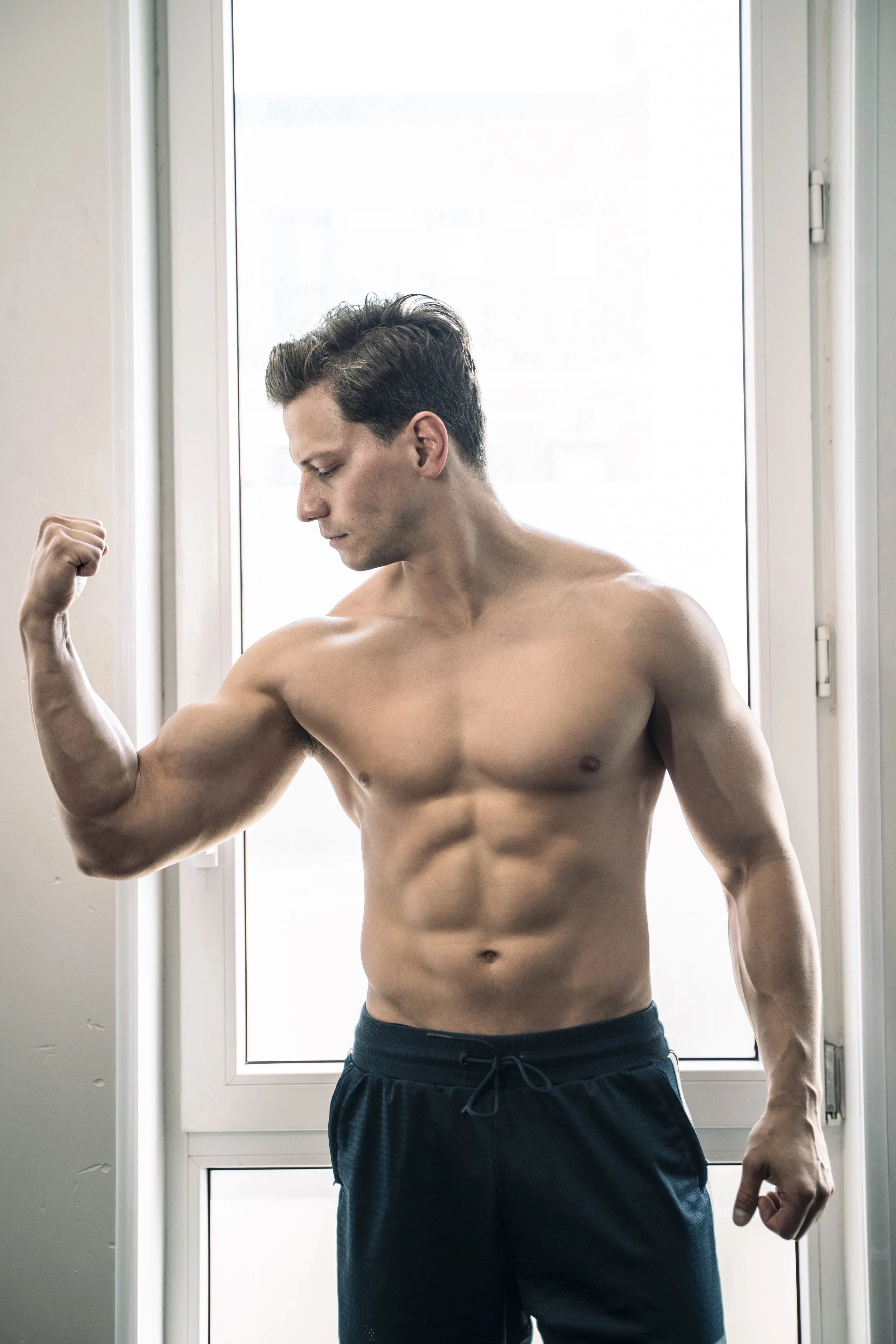 A shirtless man with muscular build flexing his right arm in front of a window.