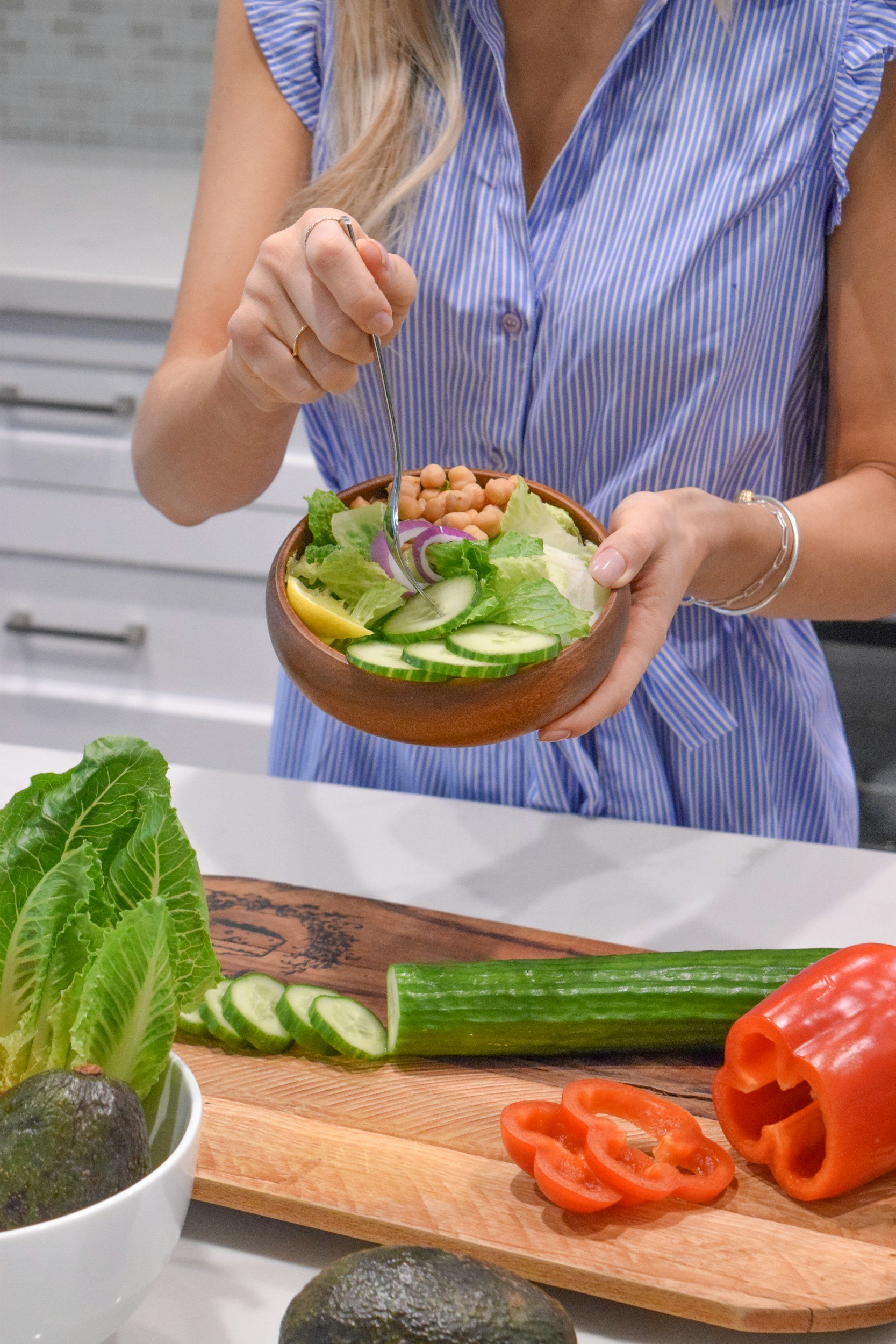 Person in blue striped shirt preparing a fresh vegetable salad with cucumbers, lettuce, onions, and chickpeas in a wooden bowl in a modern kitchen.