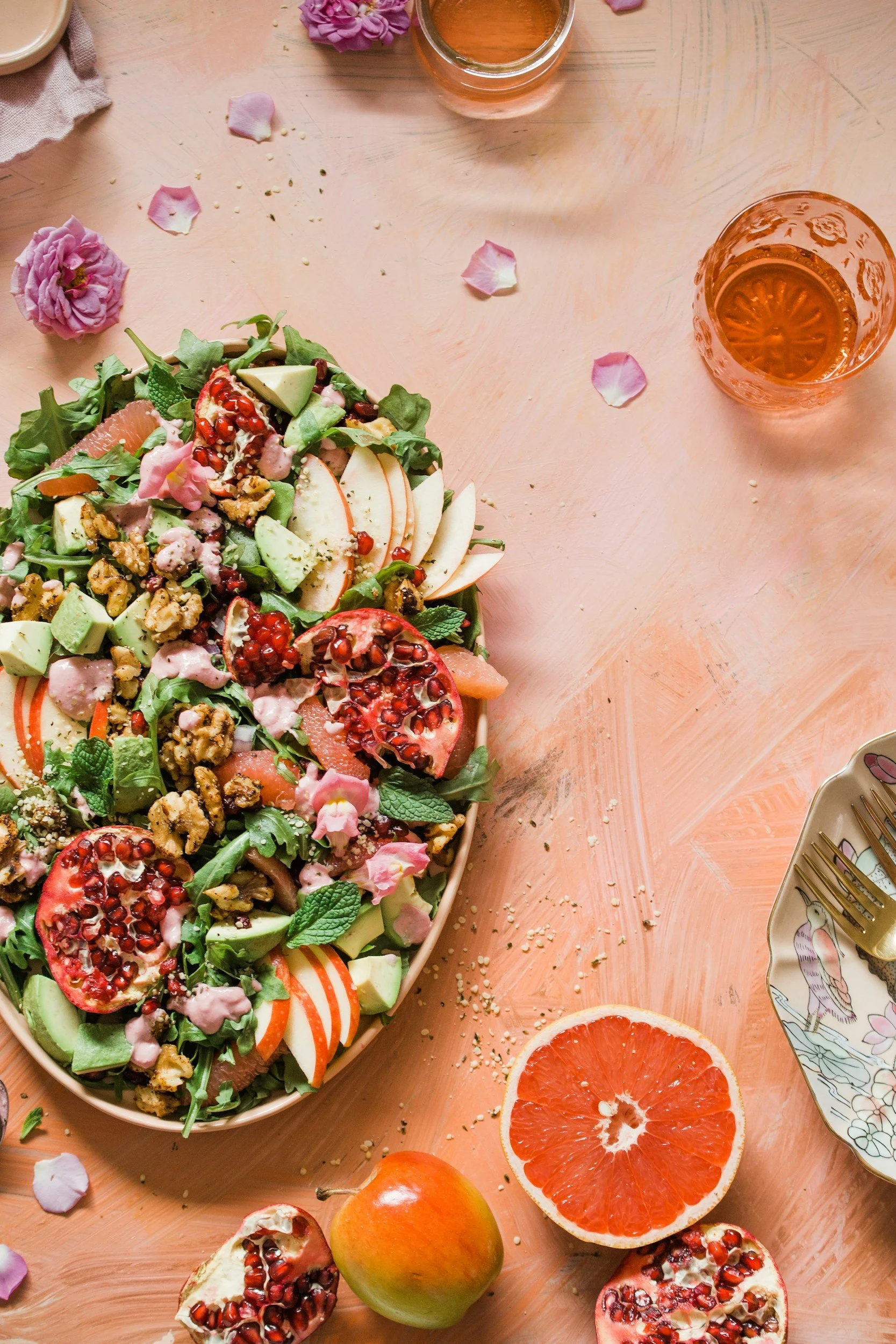 A large platter of assorted fresh fruit salad including pomegranate, apple, grapefruit, and melon on a peach-colored surface, surrounded by a halved grapefruit, an unripe apple, and pomegranate seeds, with glasses of rose-colored liquid and floral-patterned plates nearby.