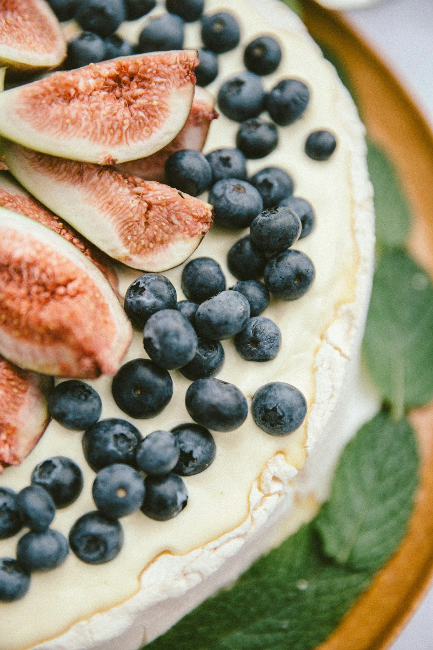 A close-up of a cheesecake topped with sliced figs, blueberries, and green leaves, on a green and brown ceramic platter.
