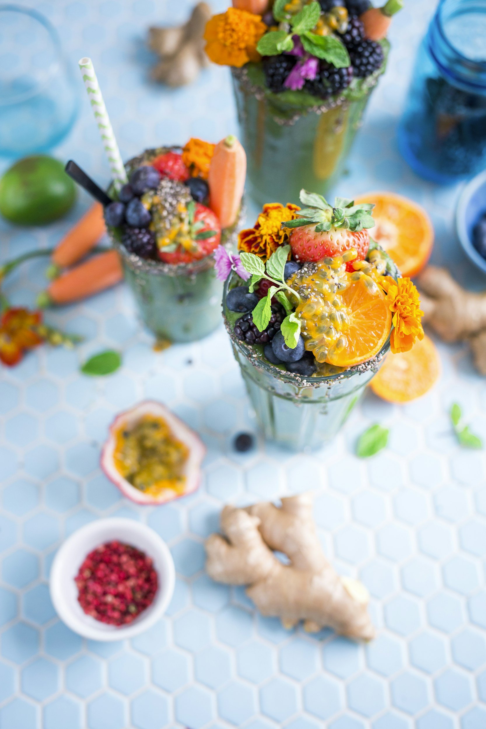 Three vibrant green smoothies topped with an assortment of fresh fruits and edible flowers, placed on a light blue tiled surface. Surrounding ingredients include half an orange, ginger root, passion fruit, berries, and small bowls of pink peppercorns and other berries.