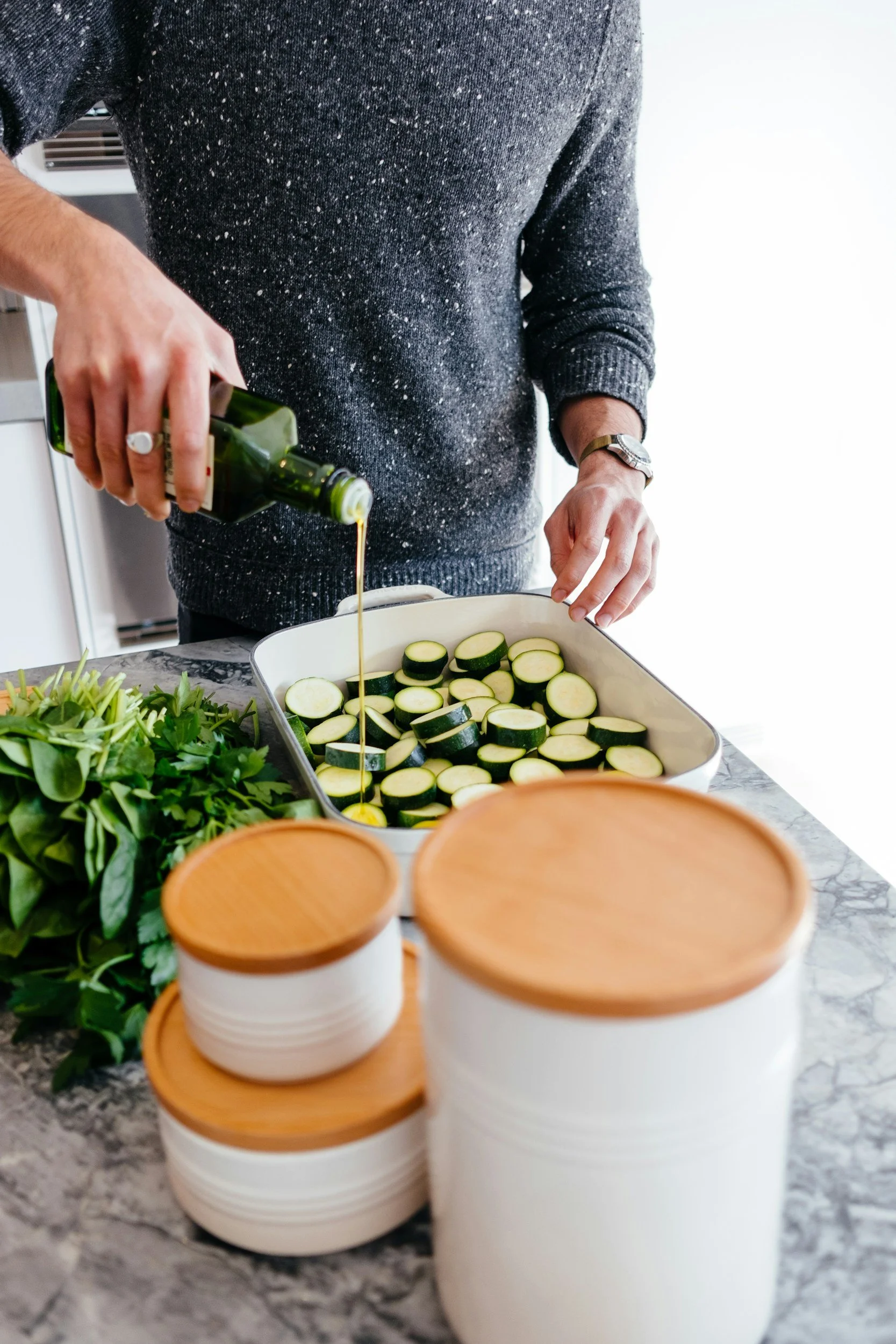 A person pouring olive oil over sliced zucchini in a baking dish on a kitchen counter, with fresh greens and stacked white bowls with wooden lids nearby.