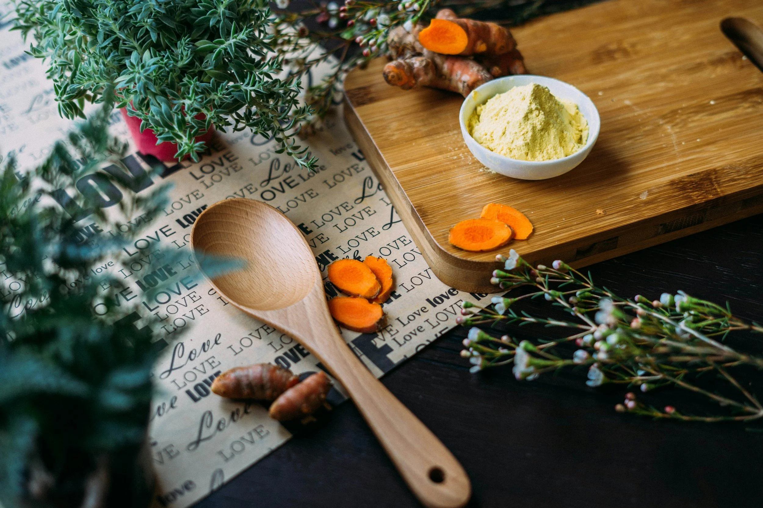 Fresh turmeric roots, sliced carrots, powdered turmeric in a bowl, greenery, and a wooden spoon on a patterned tablecloth with the word "LOVE" written repeatedly.