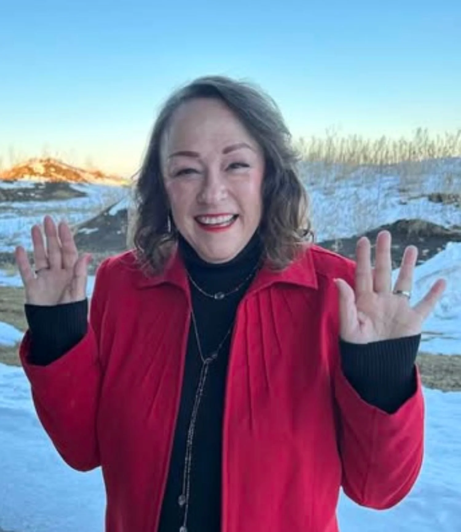 A woman in a red jacket smiling and waving outdoors in a snowy landscape with blue sky and mountains in the background.