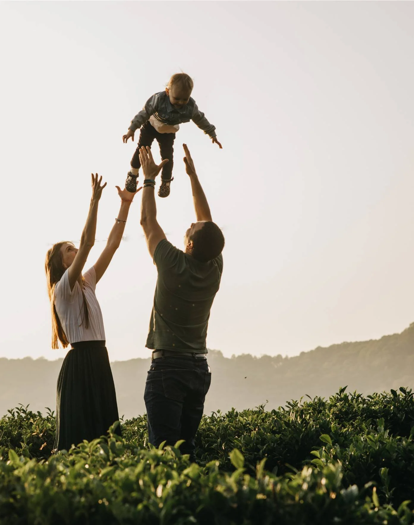 A family of three enjoying a moment outdoors, with a man and woman lifting a young child into the air amid green foliage, with hills in the background at sunset.