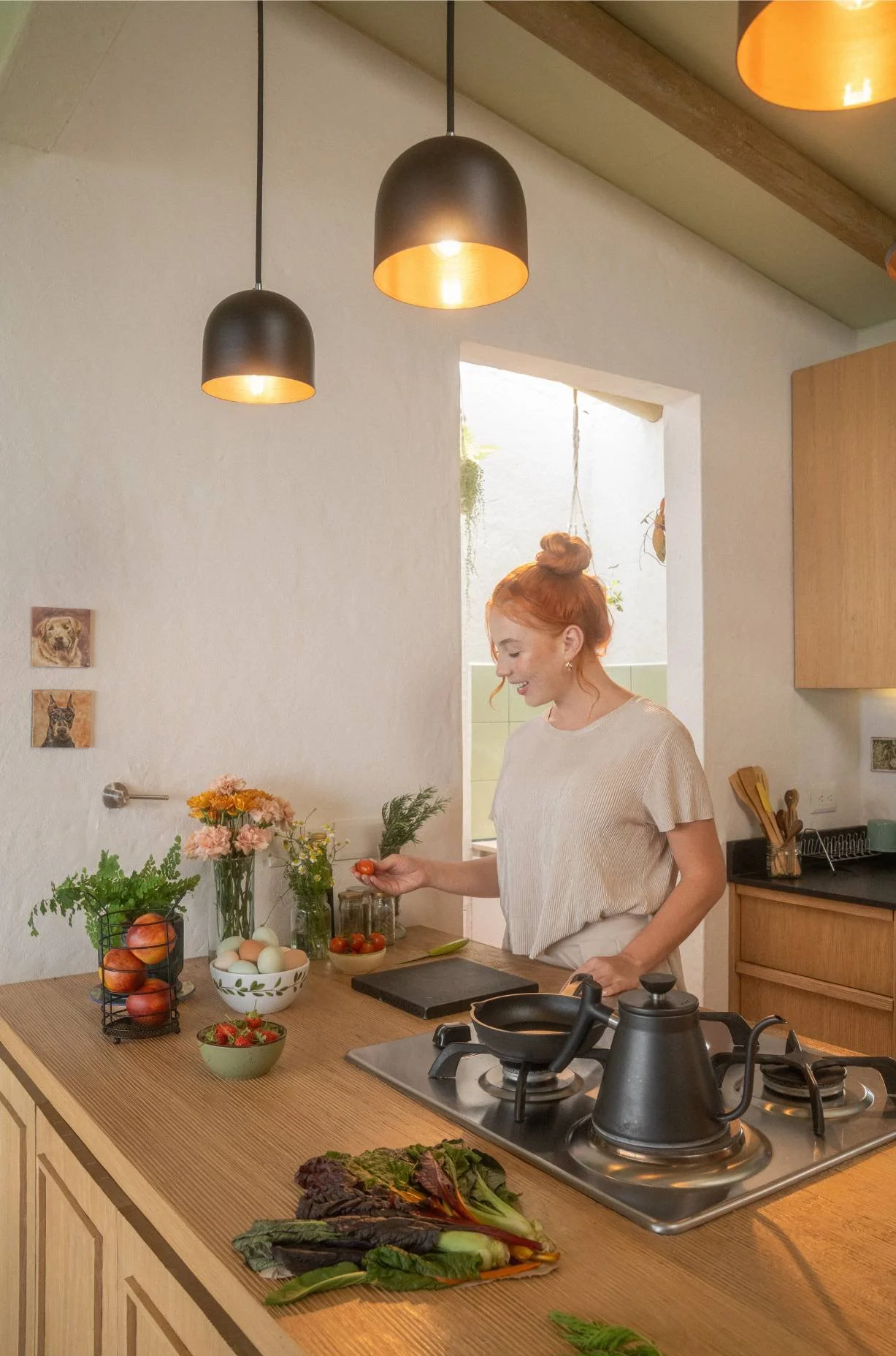 A woman in a cream-colored top preparing vegetables in a cozy kitchen with hanging black pendant lights, fresh produce, flowers, and a gas stove.
