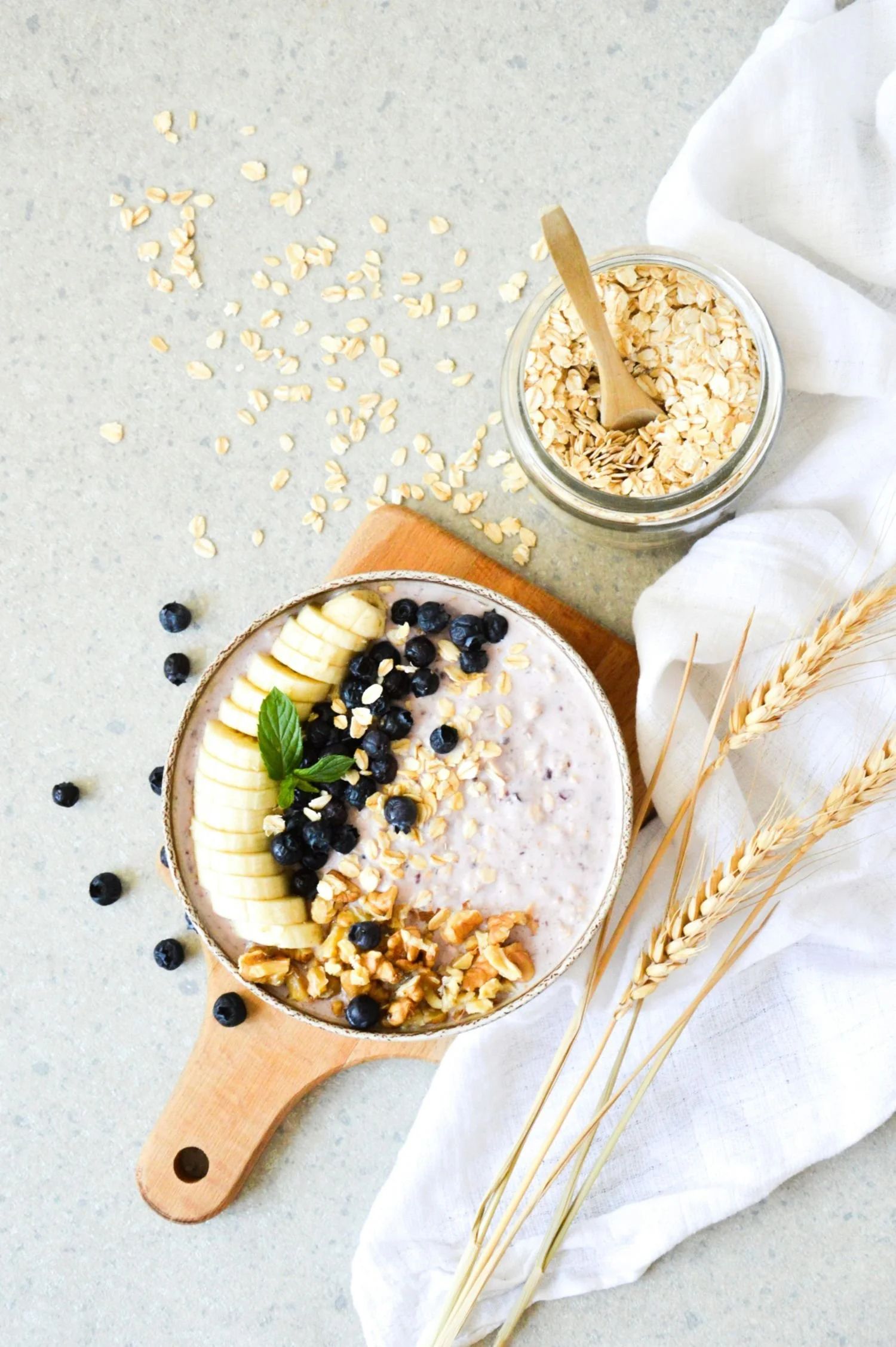 A bowl of oatmeal topped with sliced banana, blueberries, chopped nuts, and a mint leaf, with a jar of oats and wheat stalks nearby on a light surface.
