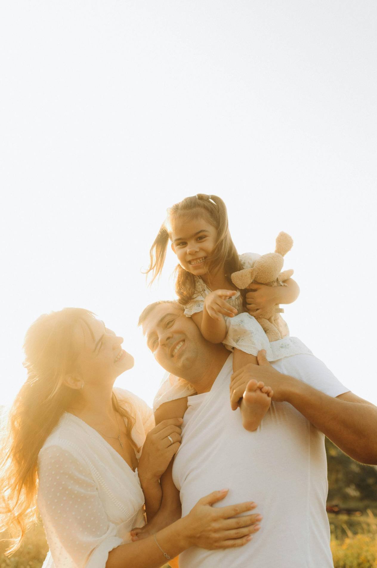 A happy family of three outdoors during sunset; a man carrying a young girl on his shoulders, and a woman standing beside them, smiling at each other.