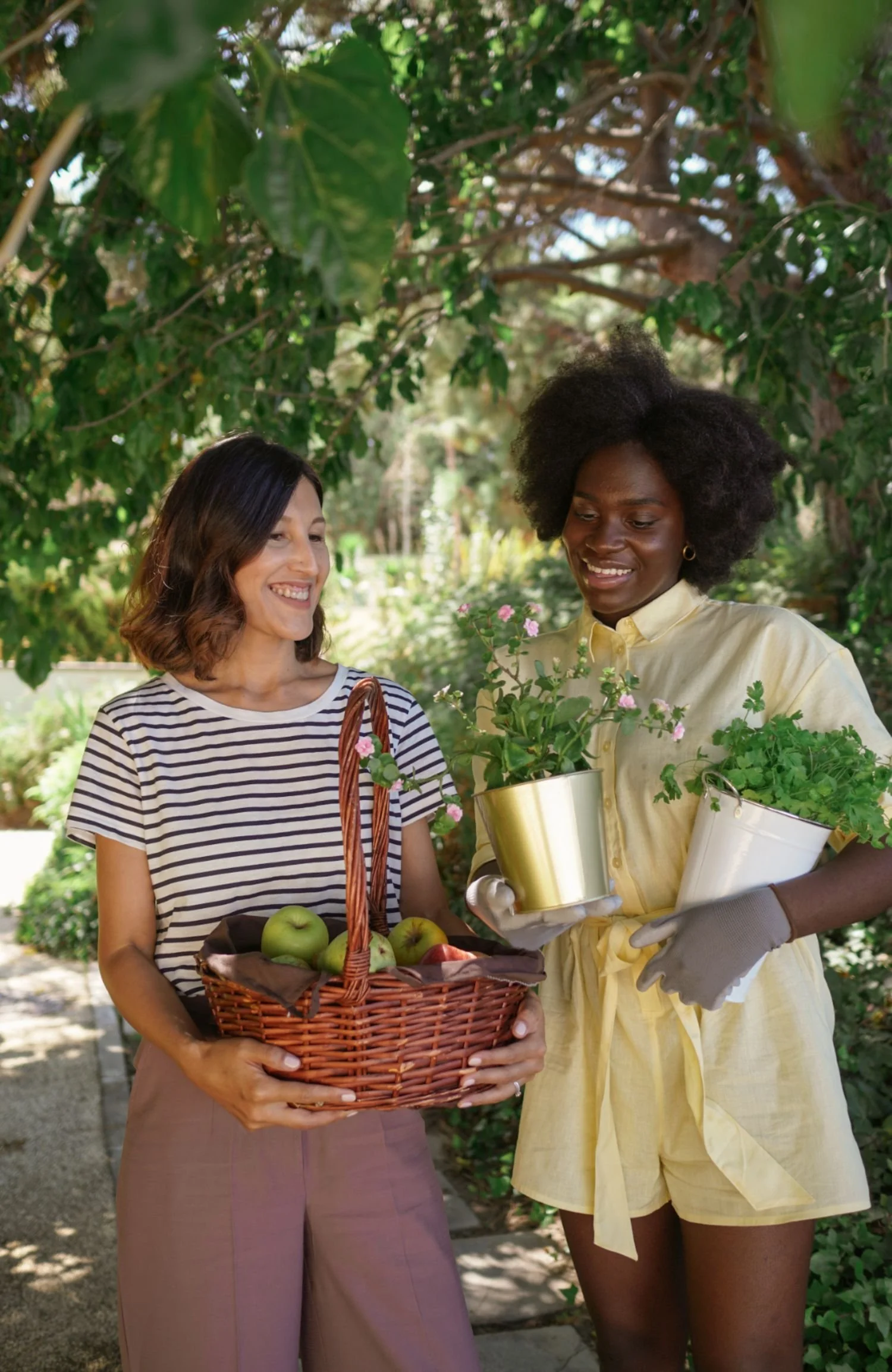 Two women in a garden, one holding a basket of apples, the other holding pots of blooming plants, smiling at each other.