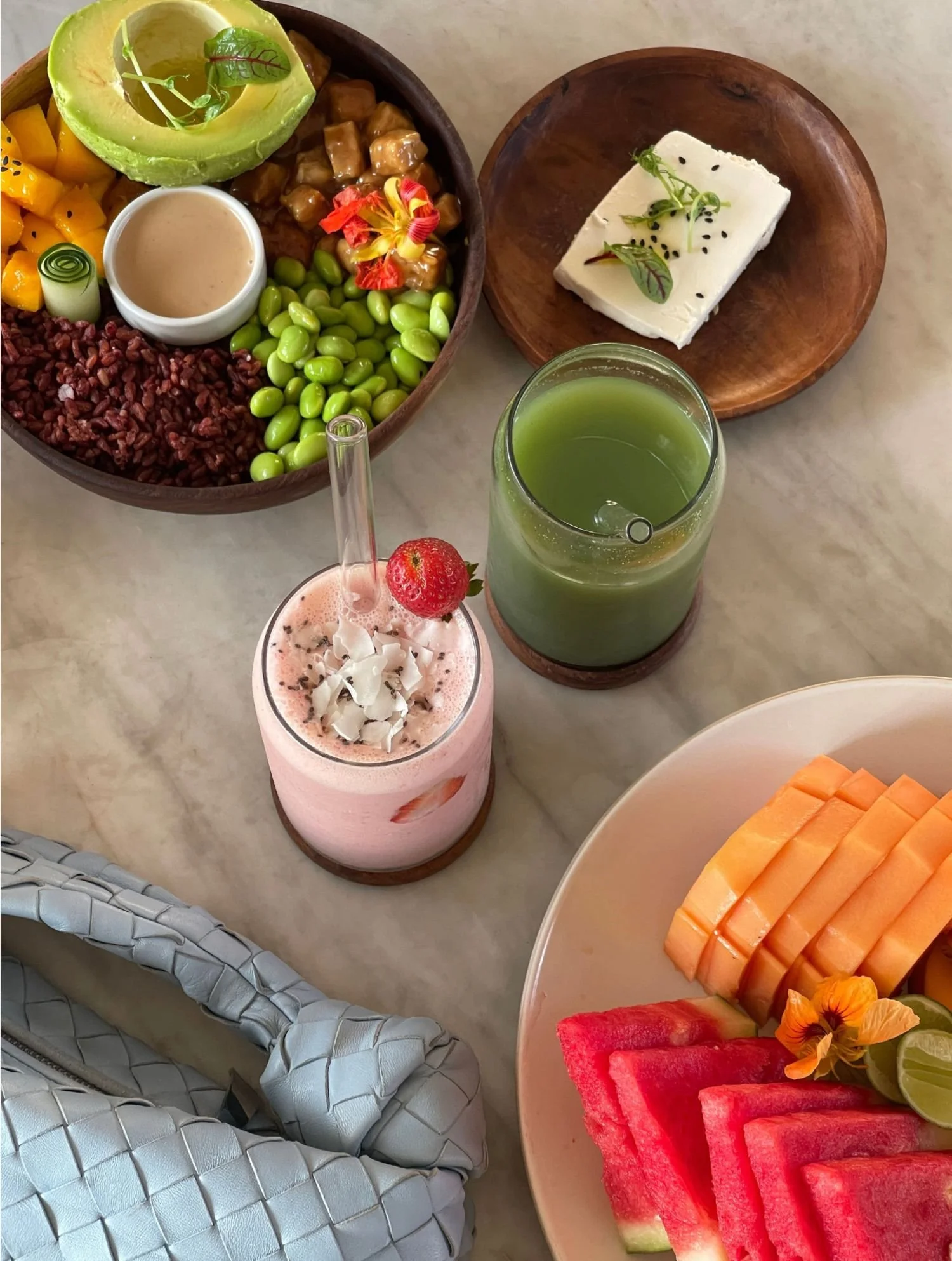 A breakfast spread including a fruit platter with watermelon, cantaloupe, and lime, a smoothie with coconut flakes and a strawberry, a green juice, a bowl of mixed fruit with mango, peaches, and edible flowers, a wooden bowl with edamame, a small plate with cheese garnished with herbs, and a large bowl with rice, avocado, chicken, peas, and vegetables.