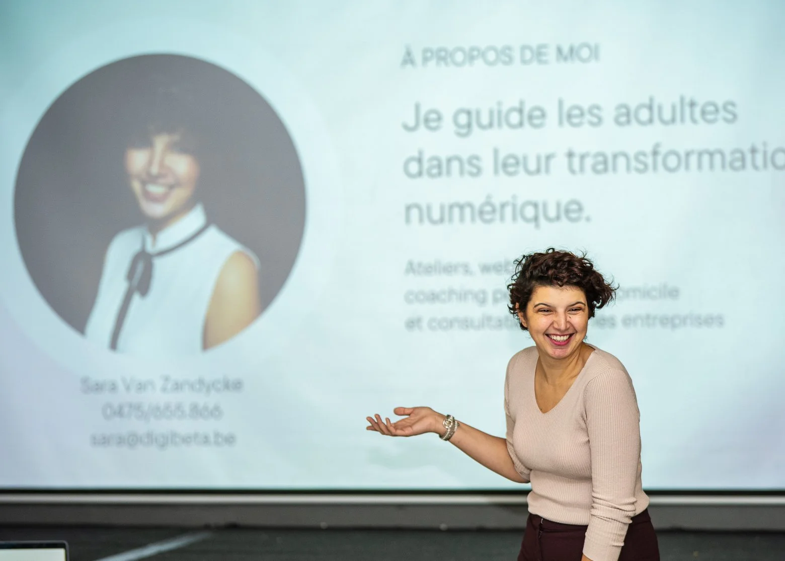 Woman smiling and gesturing towards a presentation slide featuring a photograph and biography of Sara Van Zandycke, describing her role in guiding adults in digital transformation and coaching services.