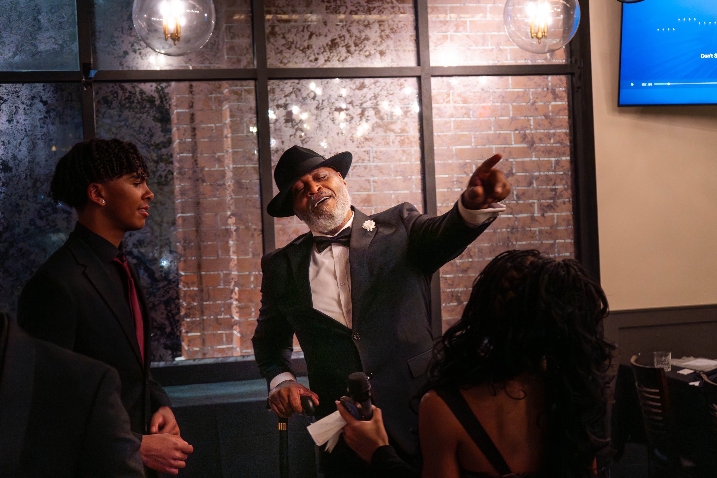 Man in suit and hat pointing while singing to family at an engagement celebration — candid event photography by DRiccardi Photography, Long Island NY.