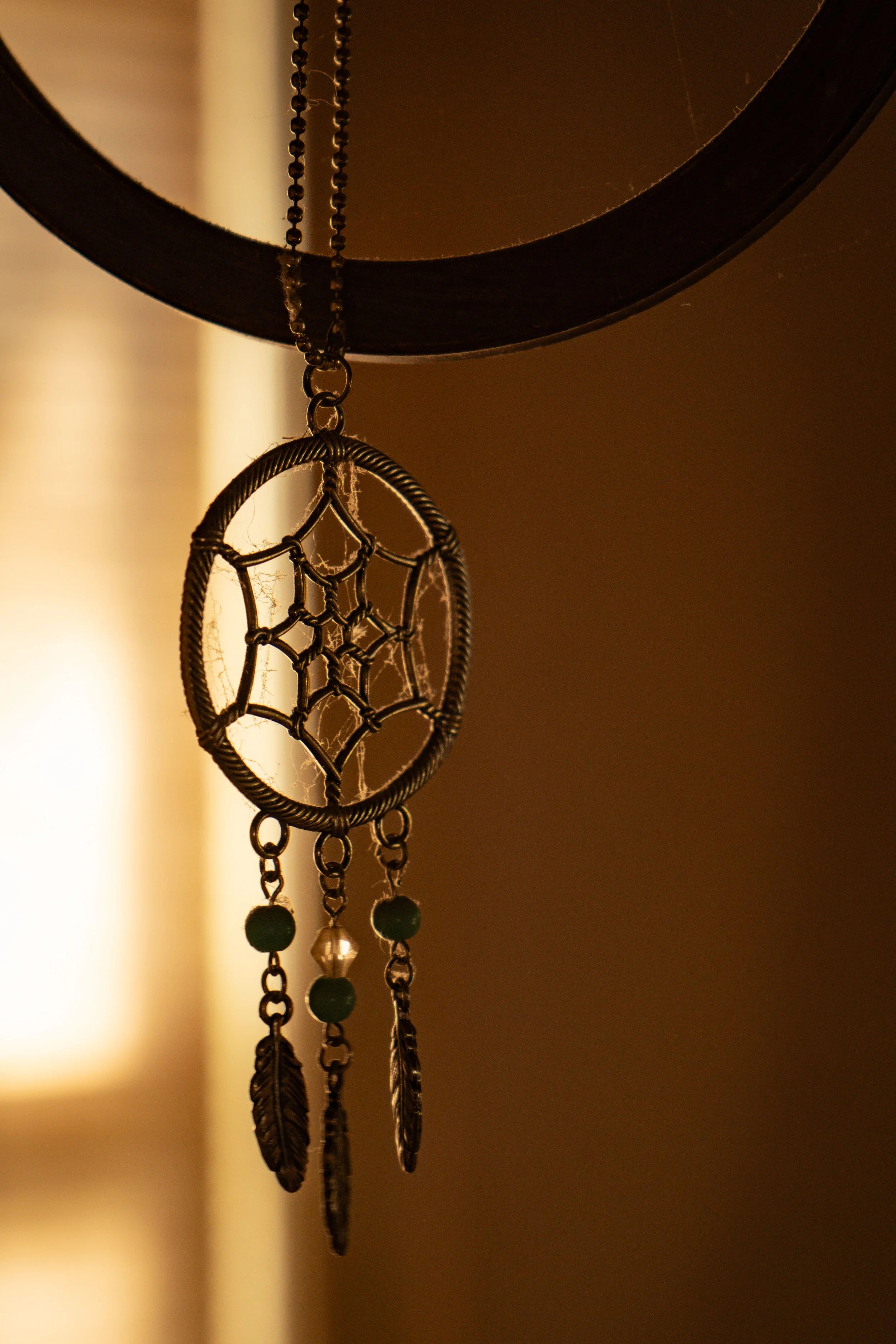Hanging dreamcatcher ornament with beads and feathers in warm window light — symbolic still-life by DRiccardi Photography.