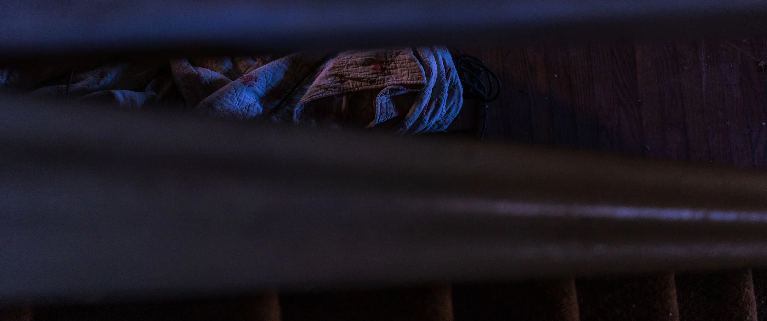 Blue-lit blanket and cord viewed through stair railing — atmospheric interior study by DRiccardi Photography, Long Island.