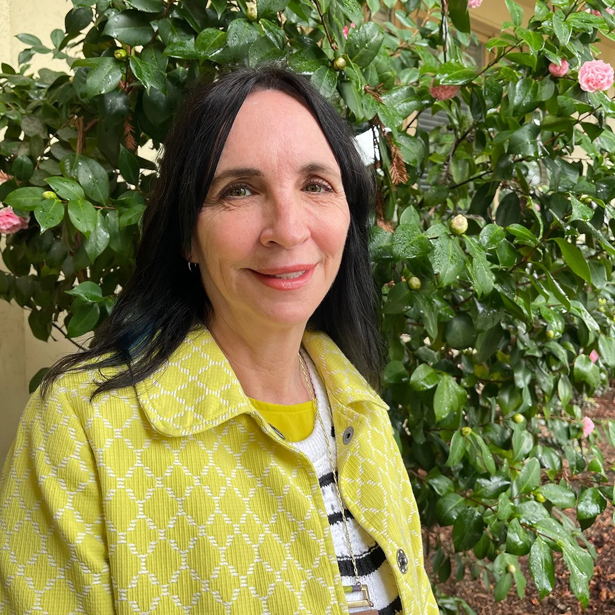 A woman with dark hair in a yellow patterned jacket standing in front of a lush green bush with pink flowers and water droplets.
