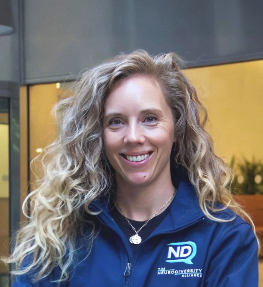 A woman with curly blonde hair smiling, wearing a navy blue jacket with the logo 'ND' and 'The Neurodiversity Alliance'