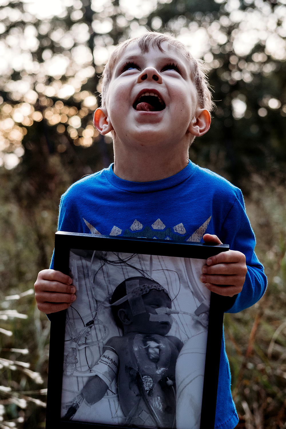 Young boy holding a framed photo of himself, looking up with open mouth and wide eyes outdoors with blurred trees in background.