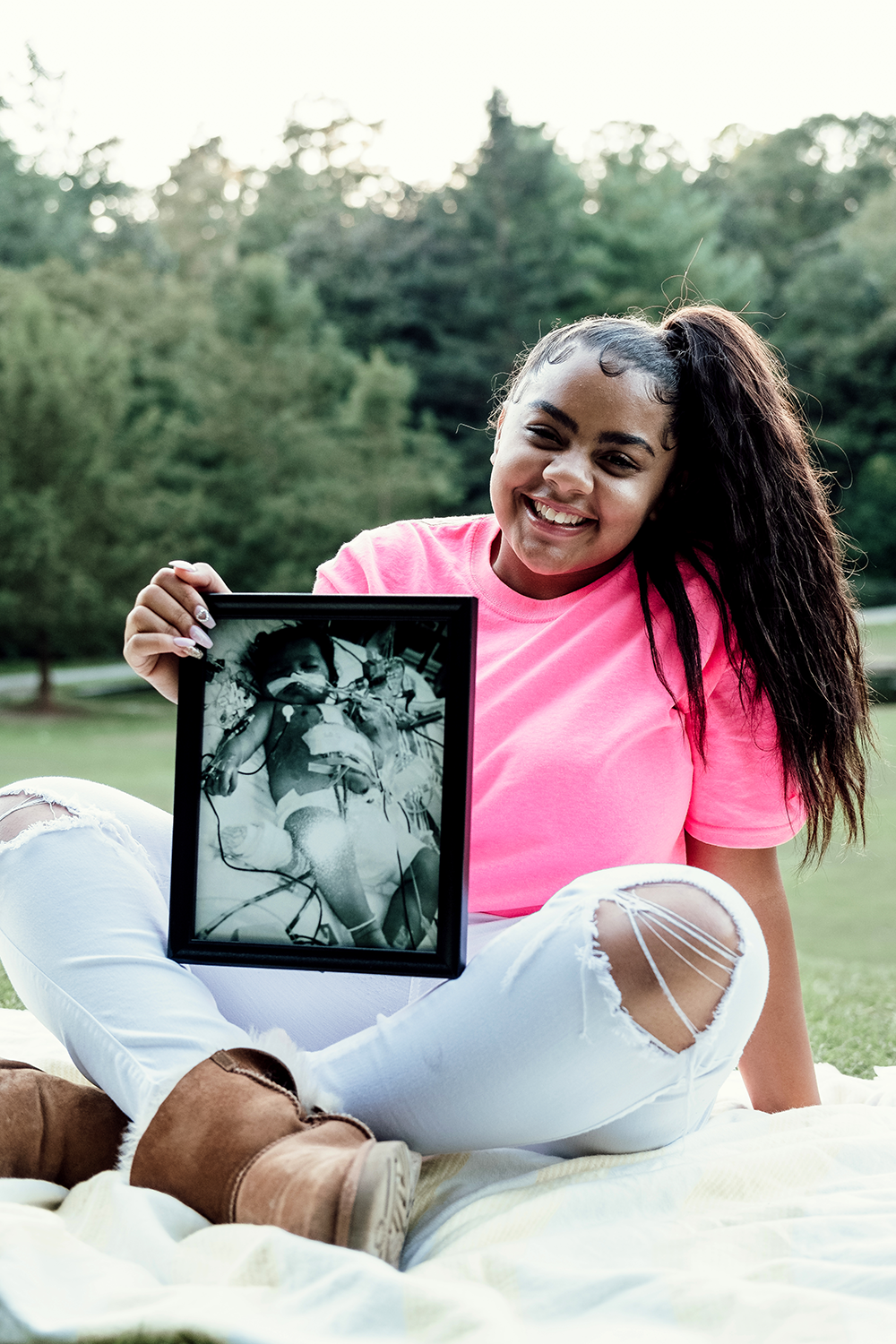 A girl sitting on grass outdoors, holding a framed black-and-white photo of herself in a hospital bed with medical wires attached, smiling and wearing a pink t-shirt and white ripped jeans.