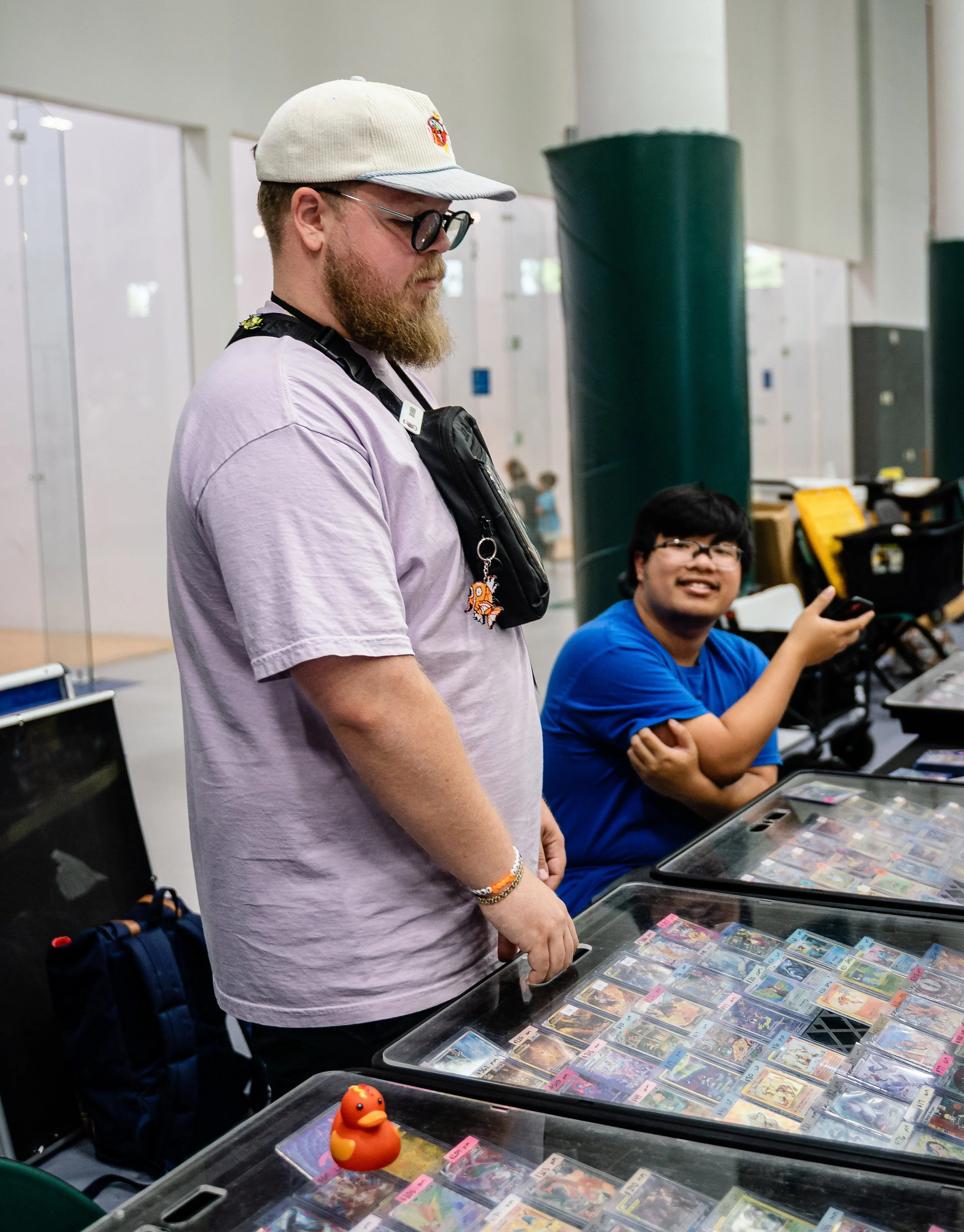A man with a beard wearing a white cap, glasses, and a purple T-shirt, standing at a table with trading cards organized in clear plastic cases, with a young boy in a blue T-shirt seated and smiling behind him, at an indoor event.