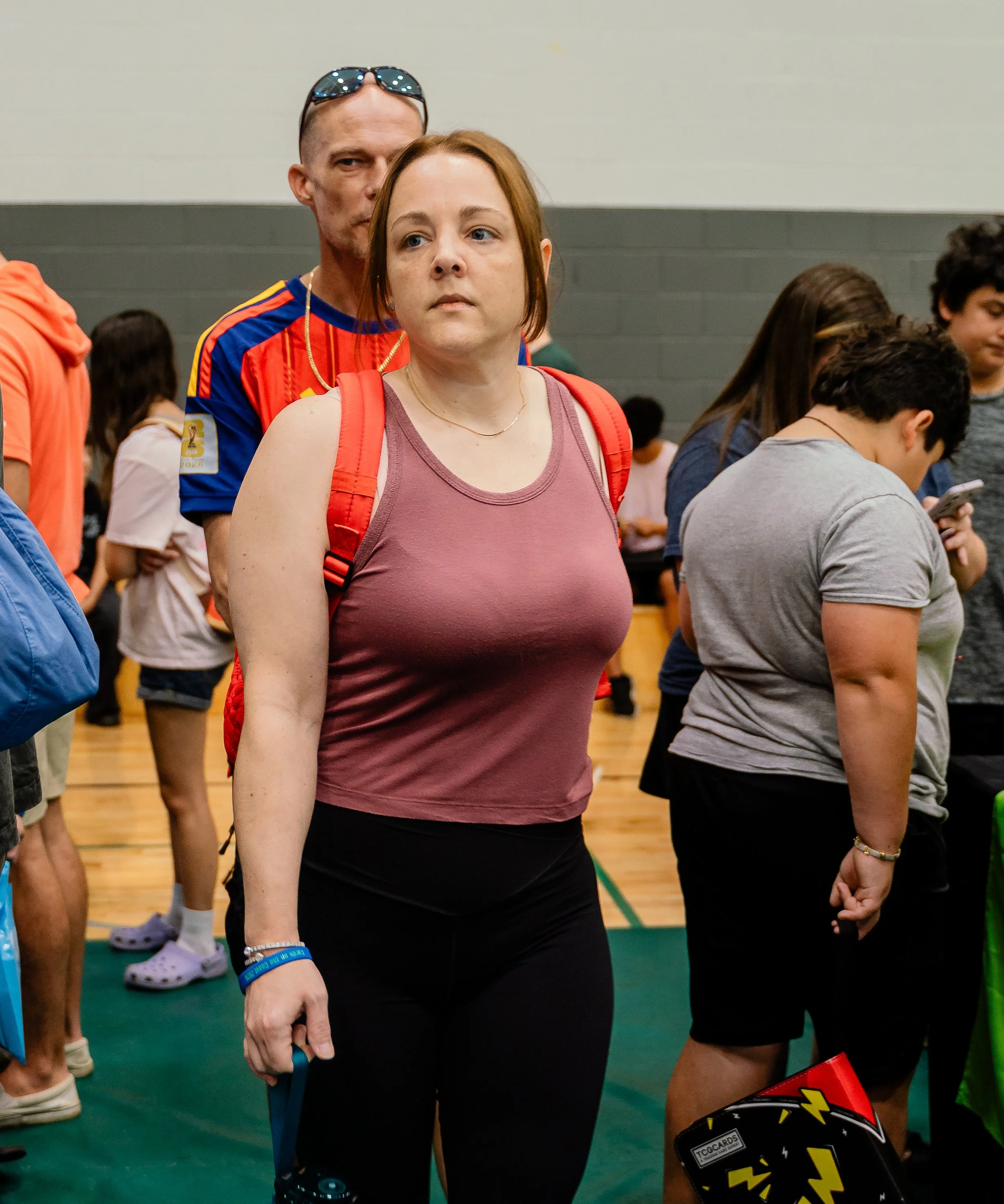 A woman with red hair wearing a purple tank top and a red backpack stands indoors, surrounded by people, some of whom are looking at their phones. The woman has a serious expression and is holding an object in her right hand. Other individuals, inclu