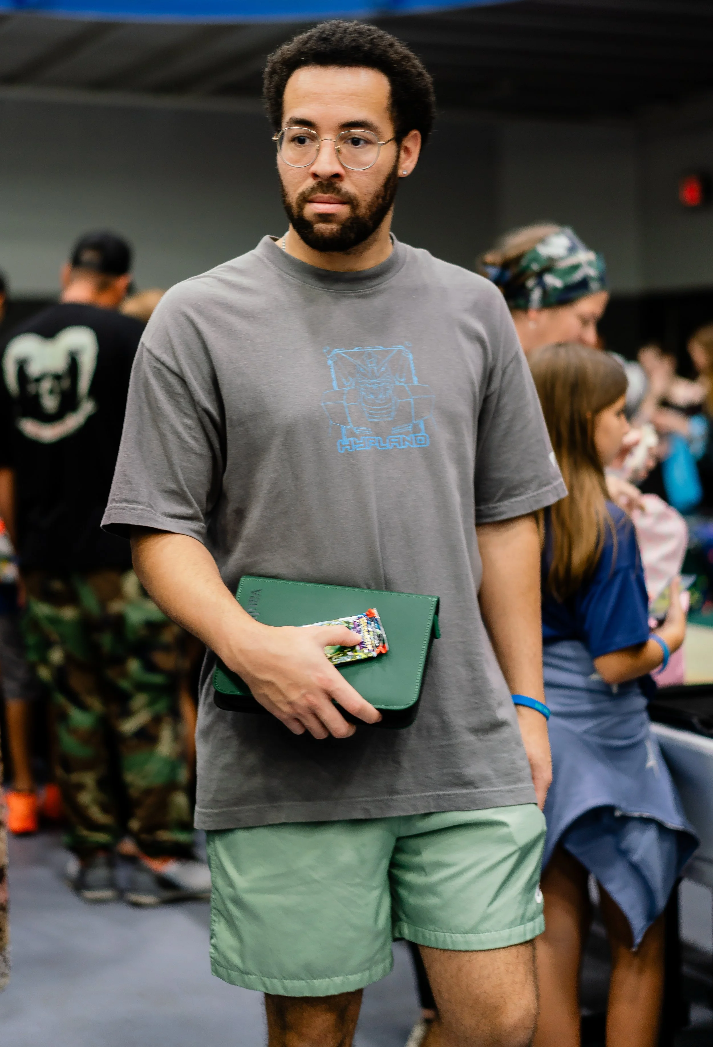 A man with glasses and a beard is standing indoors, holding a green notebook and an energy bar. He is wearing a gray t-shirt with a blue graphic that says 'HIGHLAND' and green shorts. There are other people in the background, including a woman with a