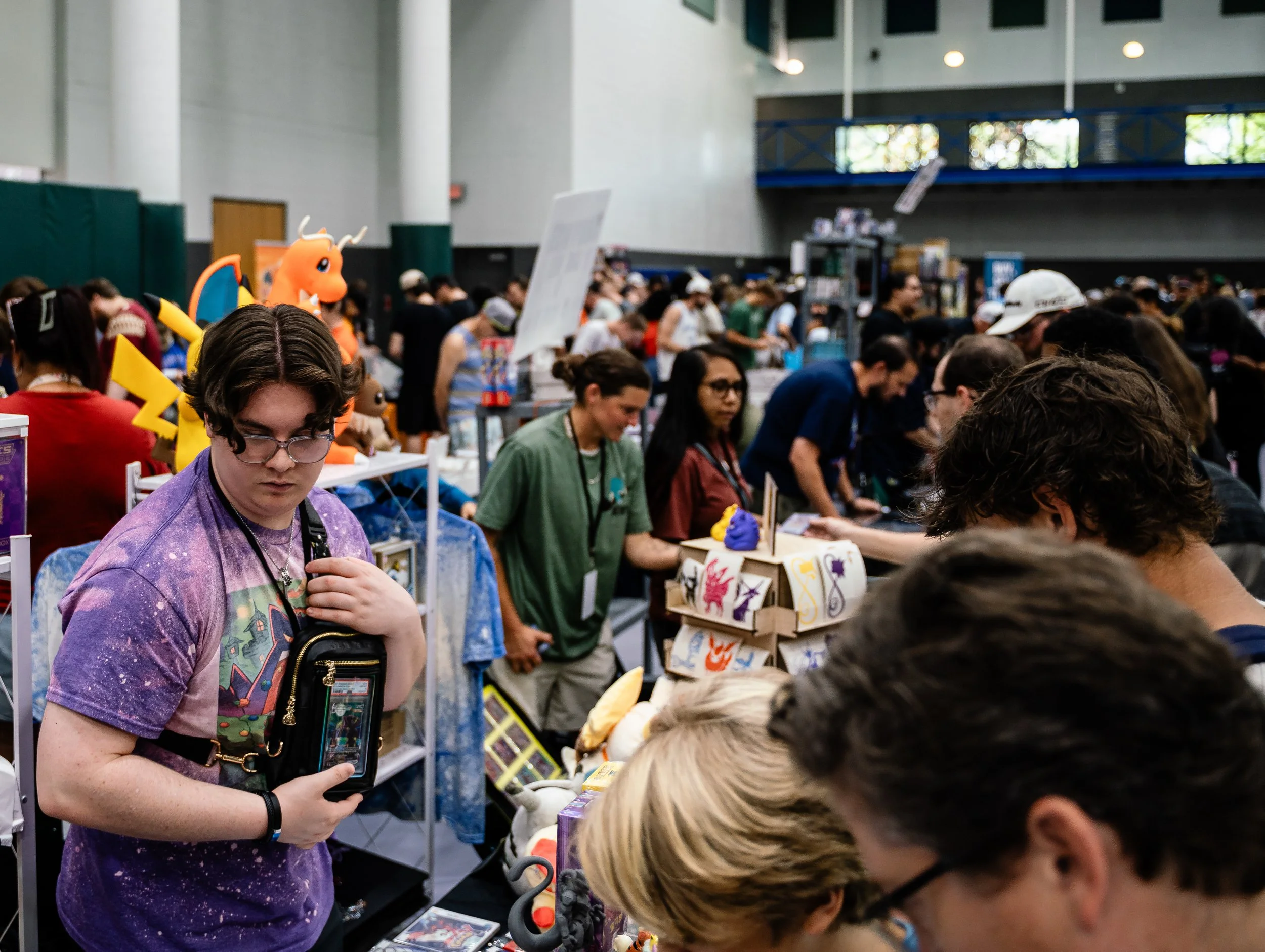 A crowded convention or event with many people browsing tables and displays, some wearing cosplay costumes or themed attire, and a person dressed as Charizard from Pokémon visible among the crowd.