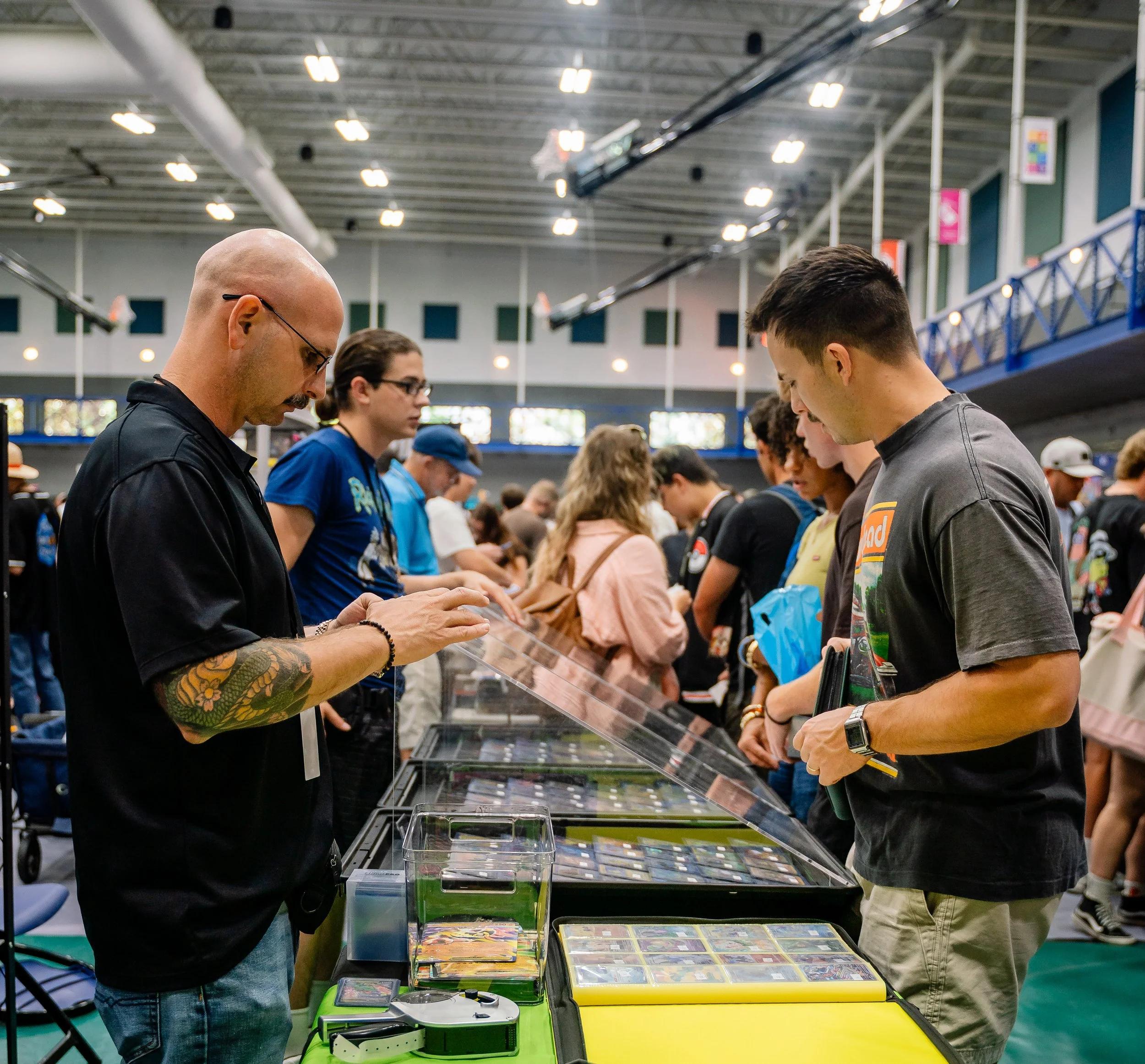 People gathered at a table browsing and purchasing trading cards at a convention or event. The background shows bright lighting and high ceilings.