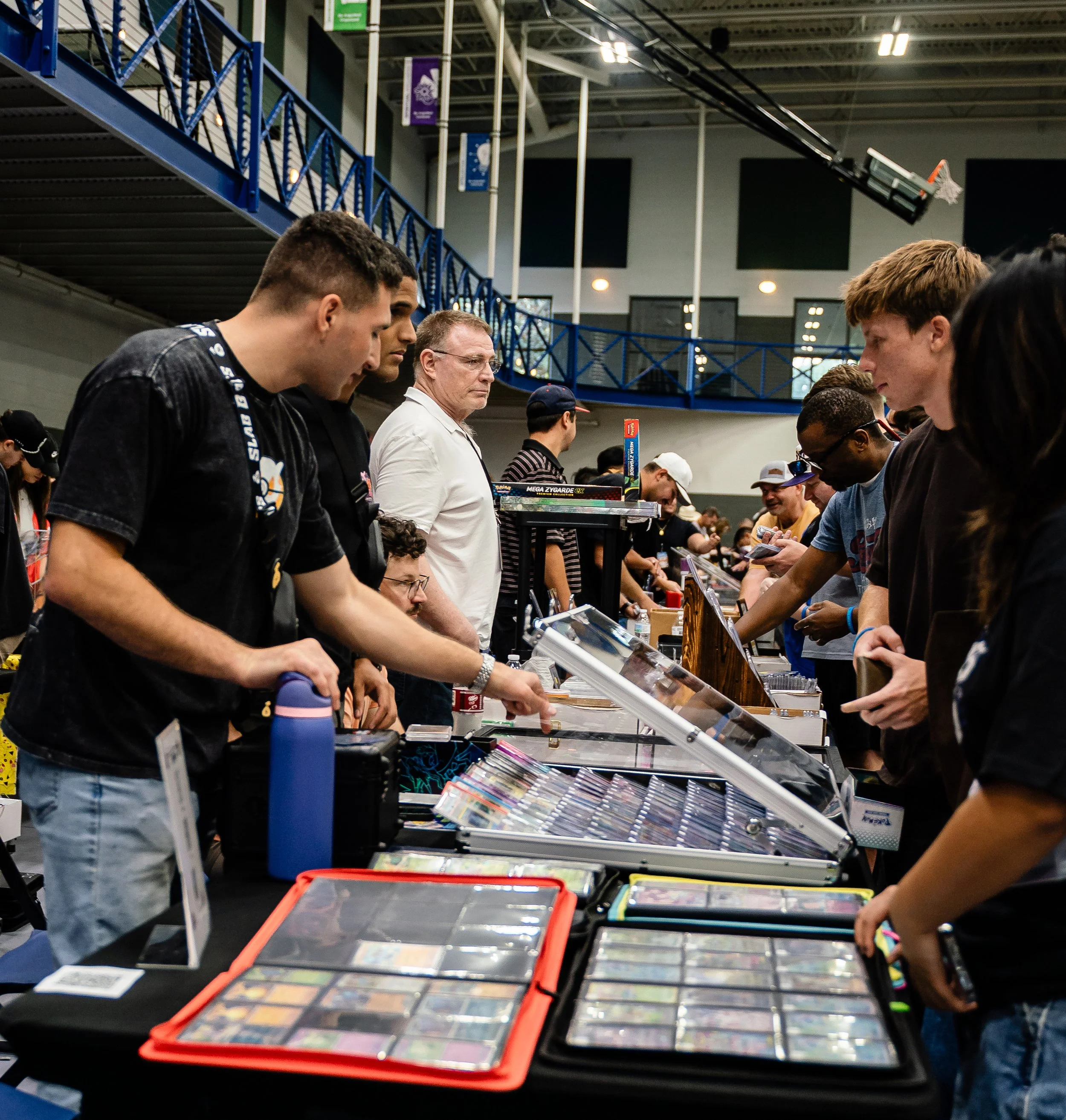 People standing at tables at a toy convention, browsing and buying collectibles and toys, with banners hanging from the ceiling in the background.
