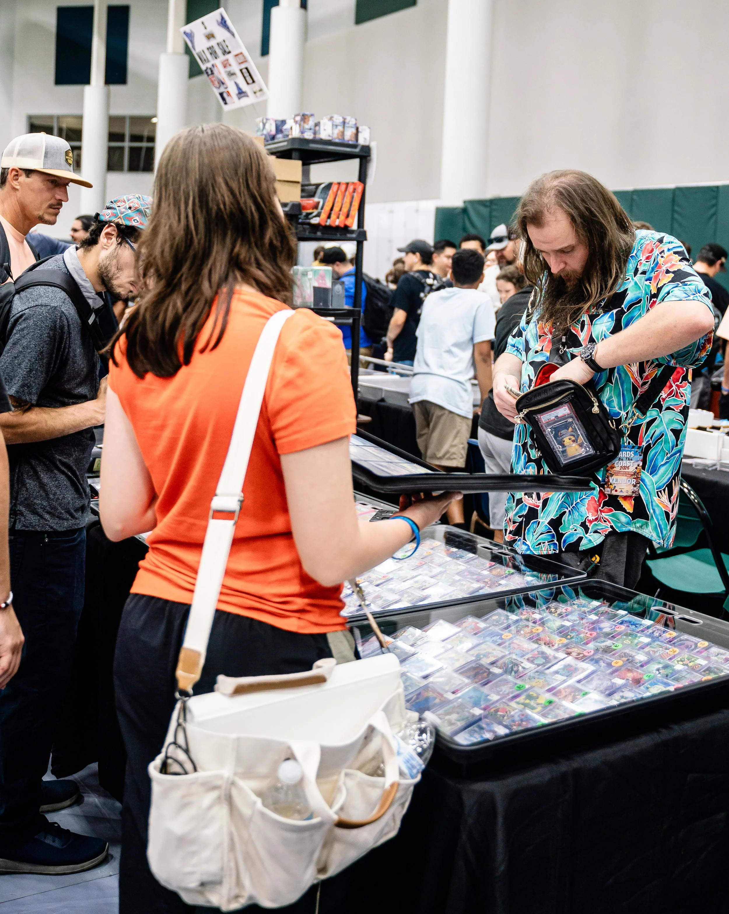 People browsing collectible trading cards at a convention or event, with a display of card packs and cases on a table.