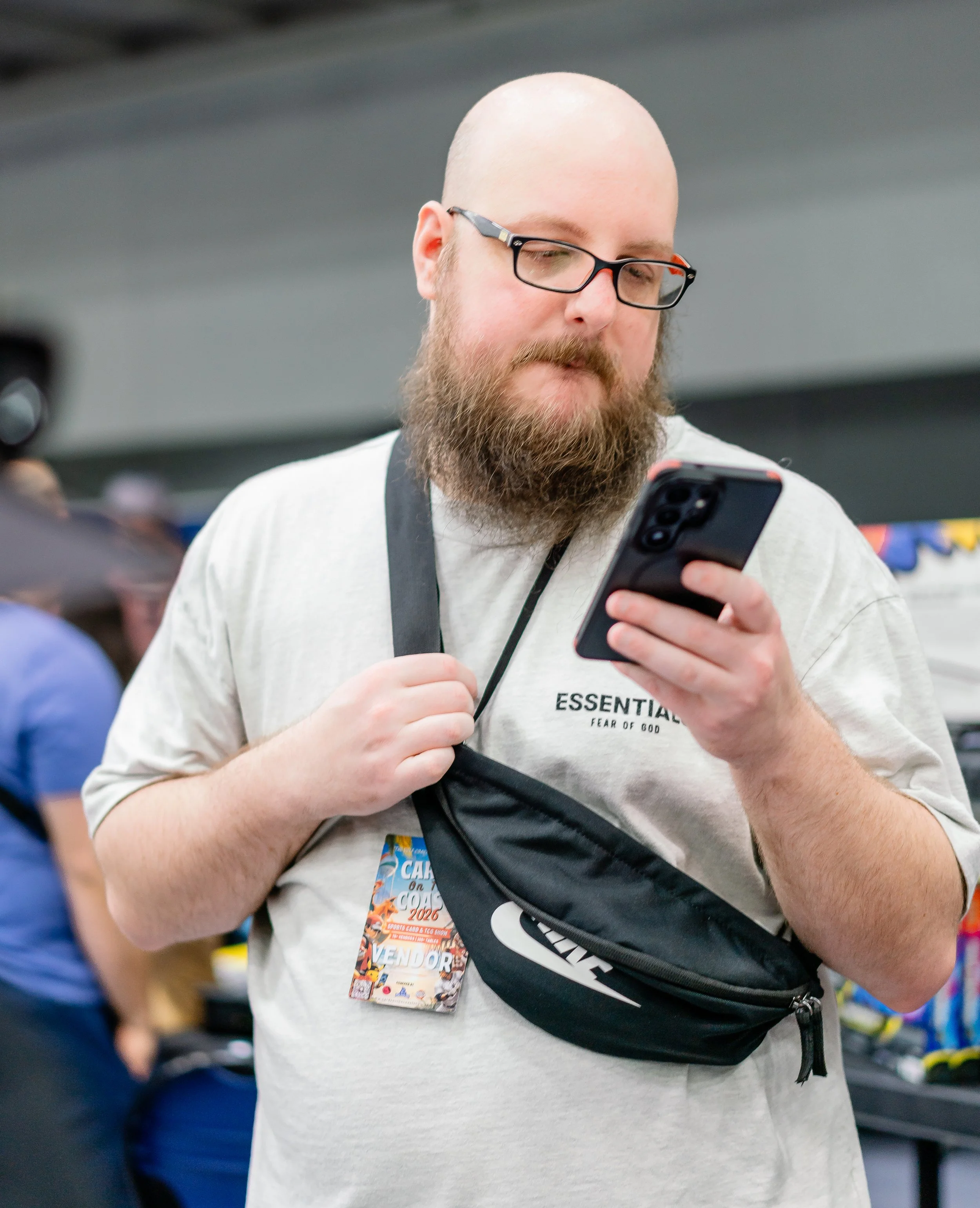 A man with a beard and glasses looking at his phone in a store, wearing a white t-shirt and a black crossbody bag.