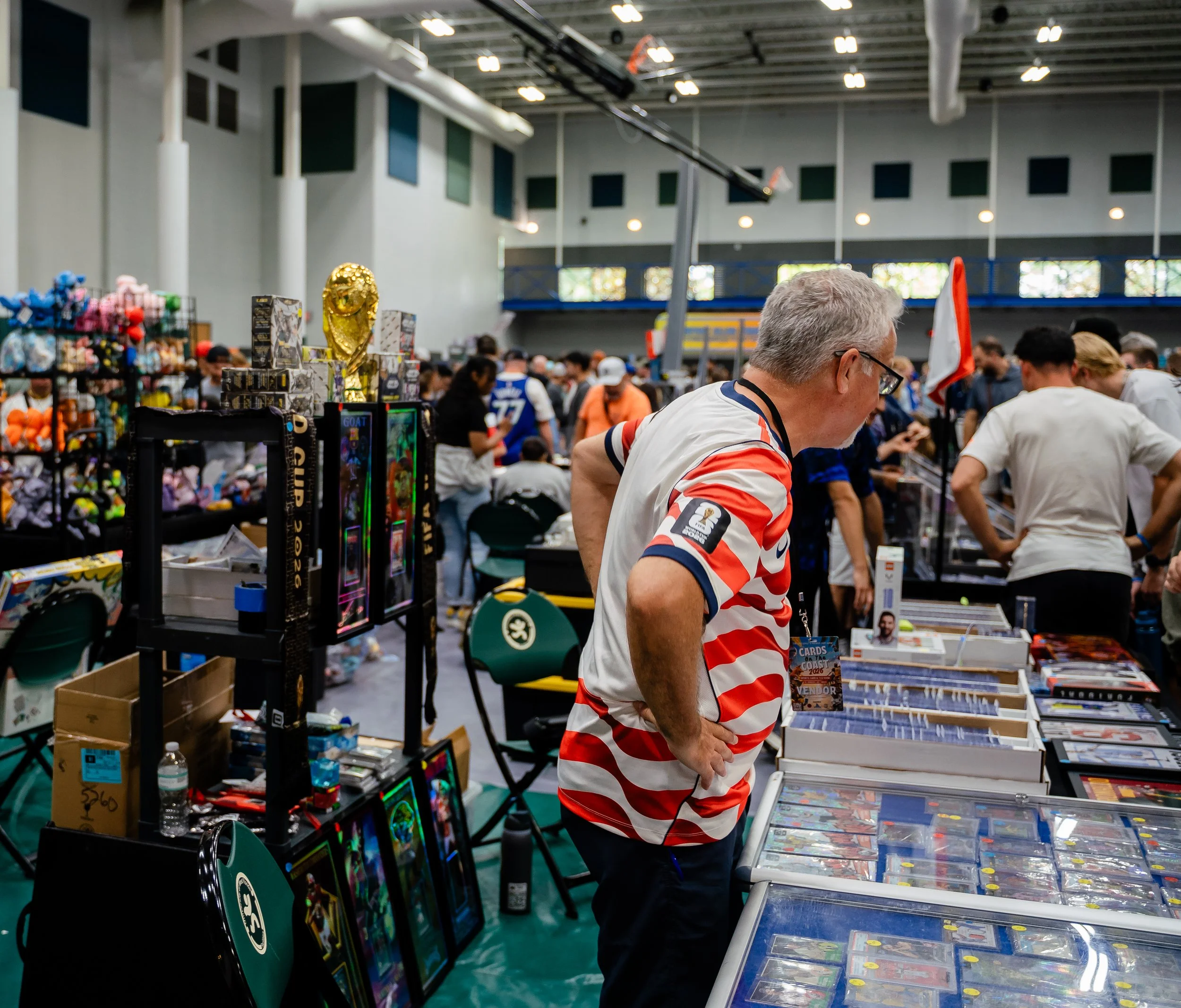 A crowded indoor market or convention with people browsing toy and collectible displays. A man in a red, white, and blue striped shirt is in the foreground. The background shows booths with toys, comics, and memorabilia, and a large gold trophy-like 