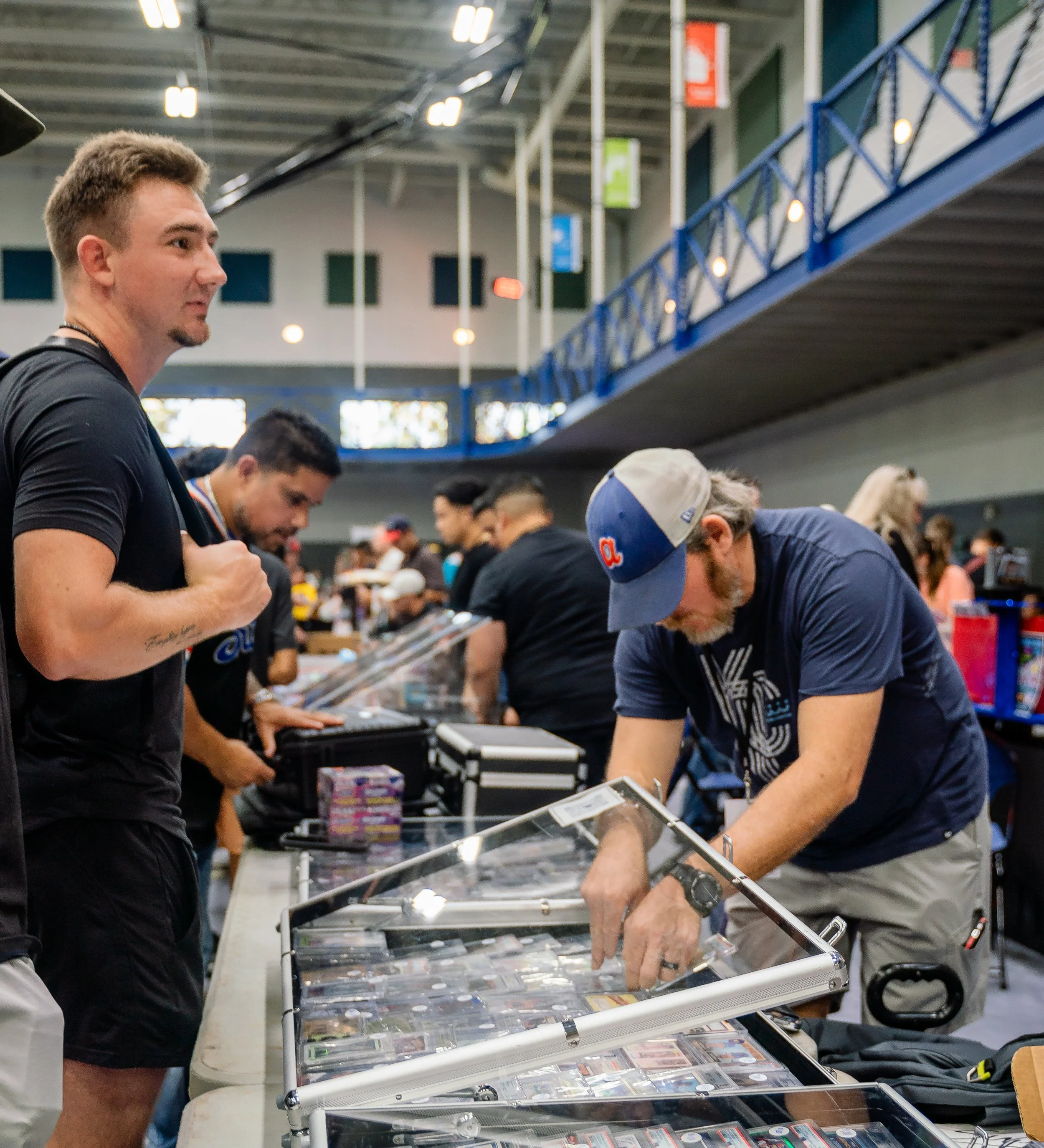 People browsing collectible cards at a convention or trade event inside a large, well-lit hall with high ceilings and colorful banners hanging from the ceiling.