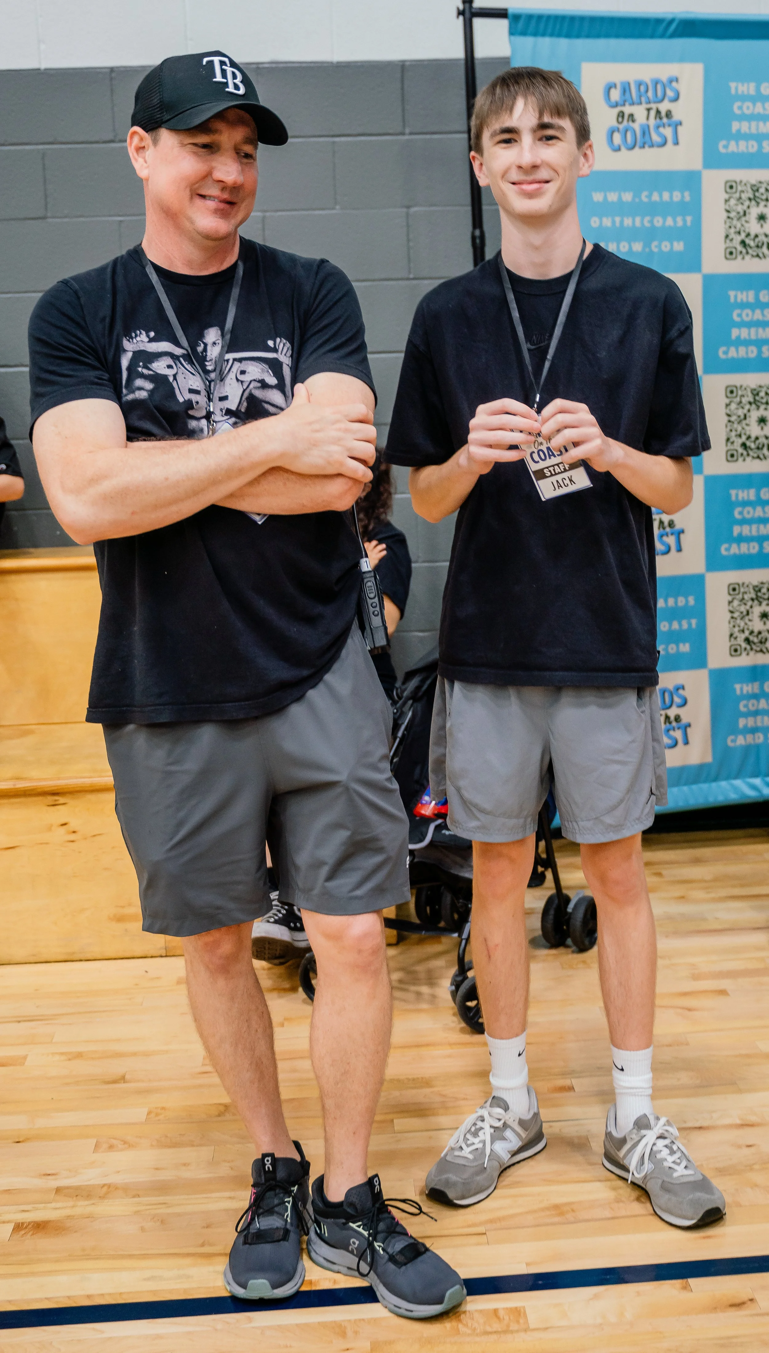Two men standing indoors, one with a Tampa Bay baseball cap and the other with a staff badge that says Jack. They are both dressed in casual athletic clothing, wearing shorts and sneakers, and smiling. Behind them is a promotional banner that reads '