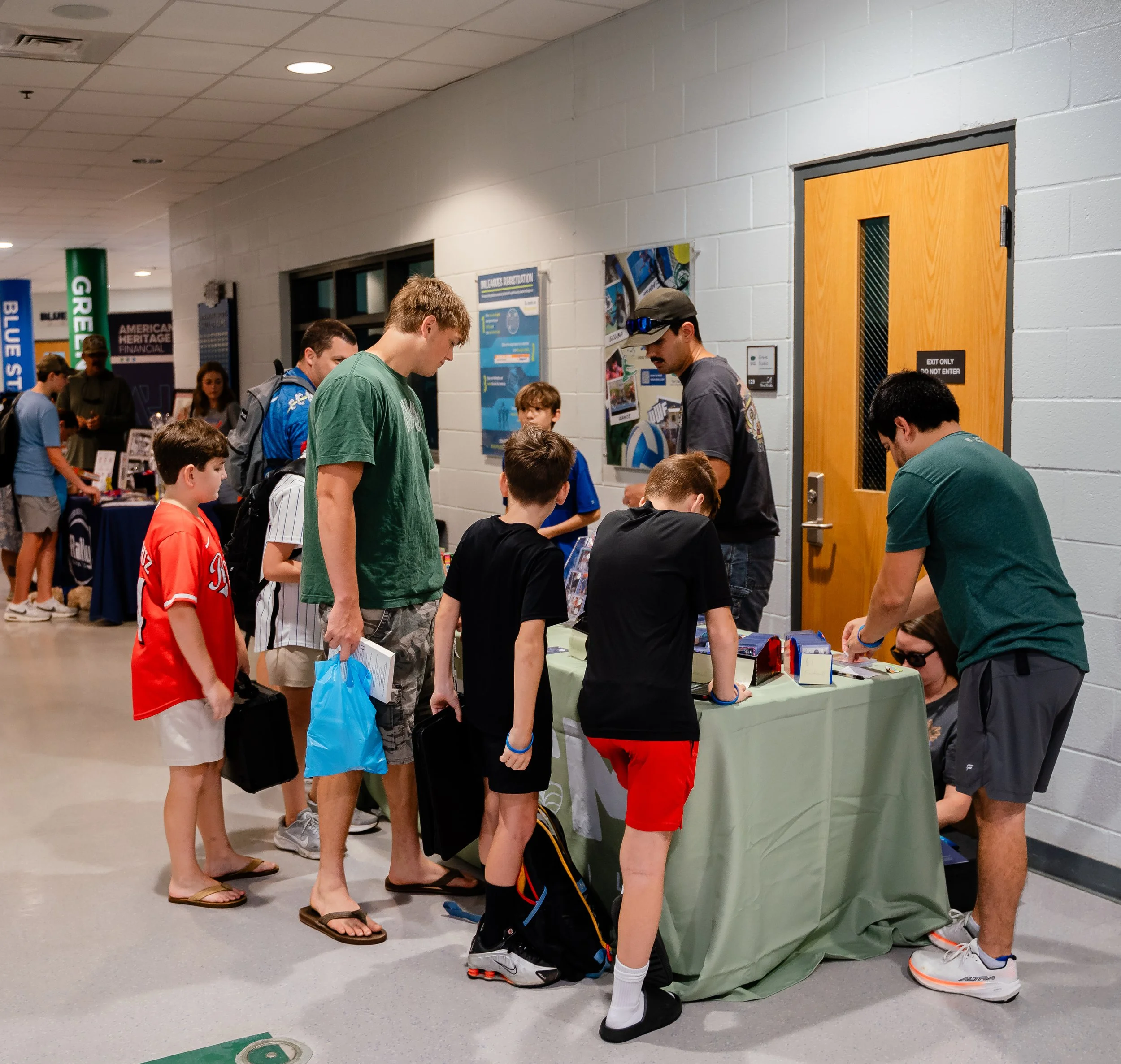 Group of people, including children and adults, standing in line at an indoor event, with some engaging with a table or booth against a light gray wall, near a wooden door with a small window and a sign that reads 'EXIT ONLY. DO NOT ENTER'.