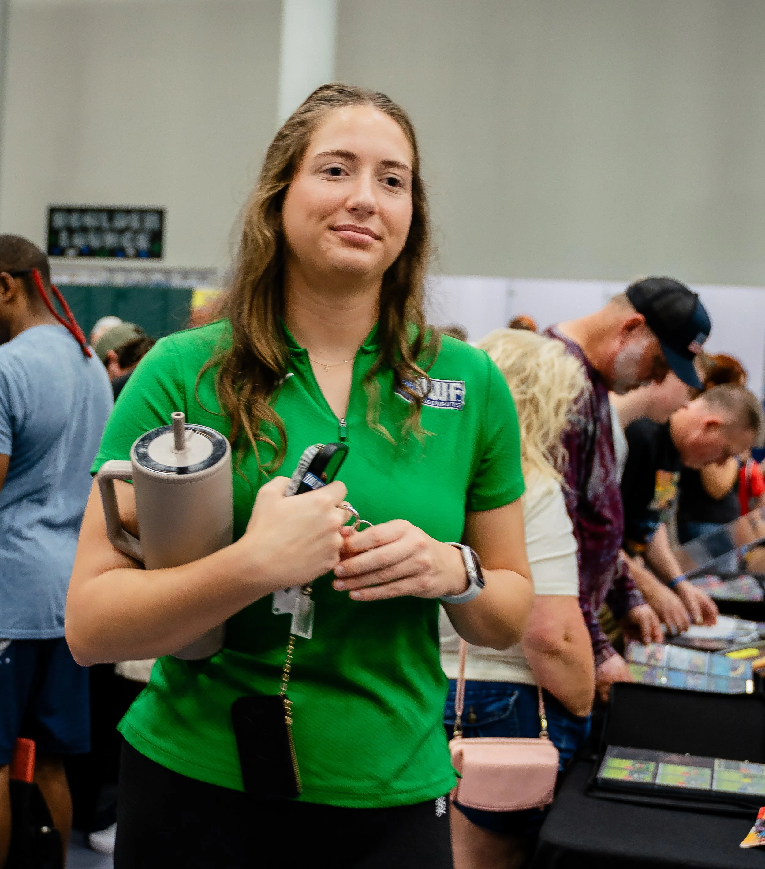 A young woman with long brown hair wearing a green shirt stands in a crowded room, holding a cup with a straw and a phone. She appears to be at an event with other people, some looking at items on a table.