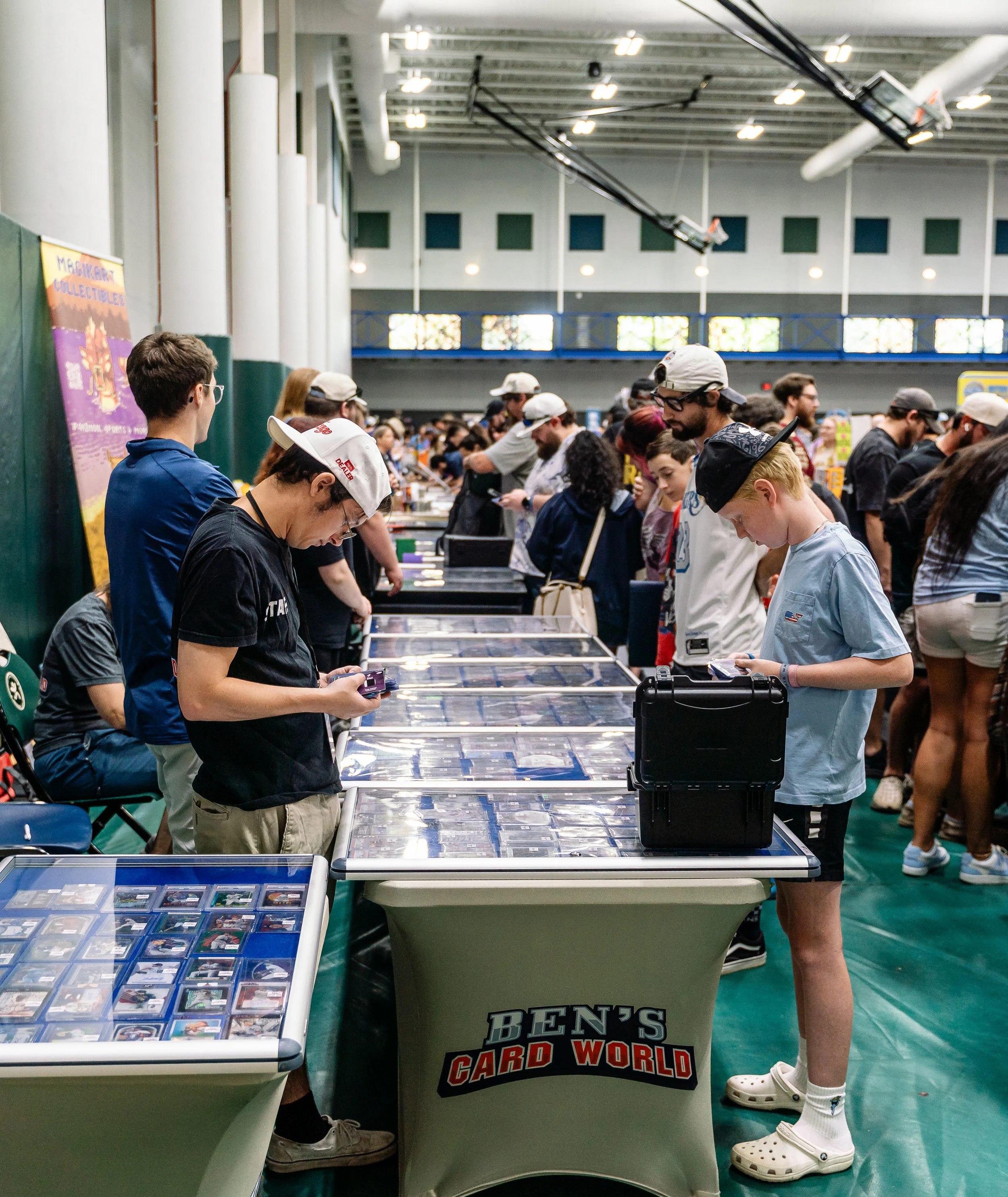 People browsing trading cards at a card show, with tables displaying collectible cards and a Ben's Card World sign on one of the tables.