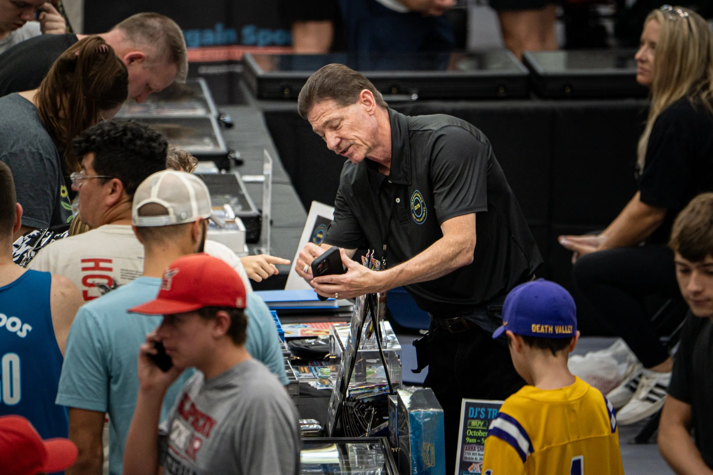 People gathered around a table at a convention or event, browsing merchandise or collectibles, with one man showing something on his phone to a group of fans.