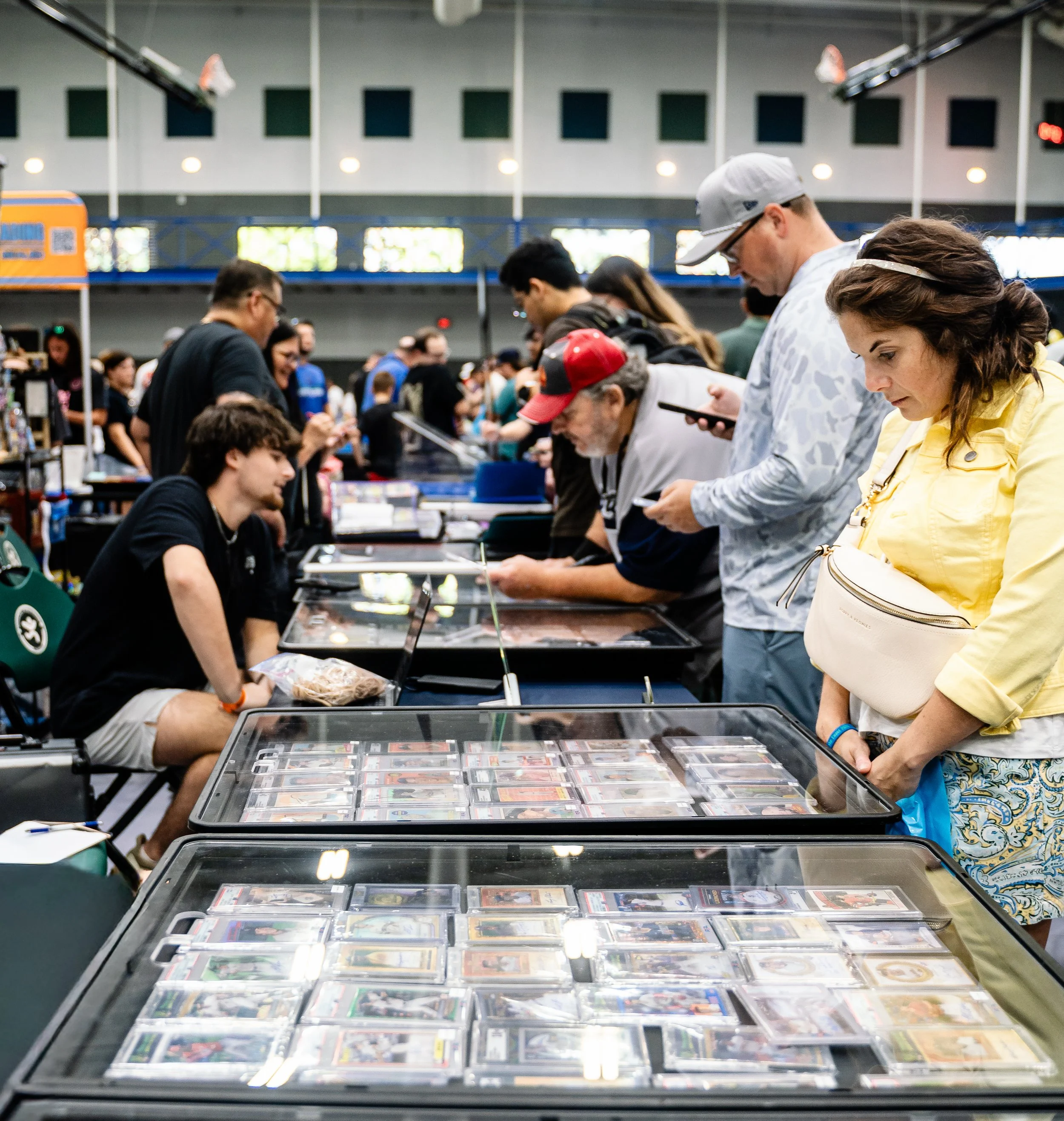 People browsing card games and collectibles at an indoor convention or event. The scene includes a long table with trays of trading cards, and attendees are focused on examining them.